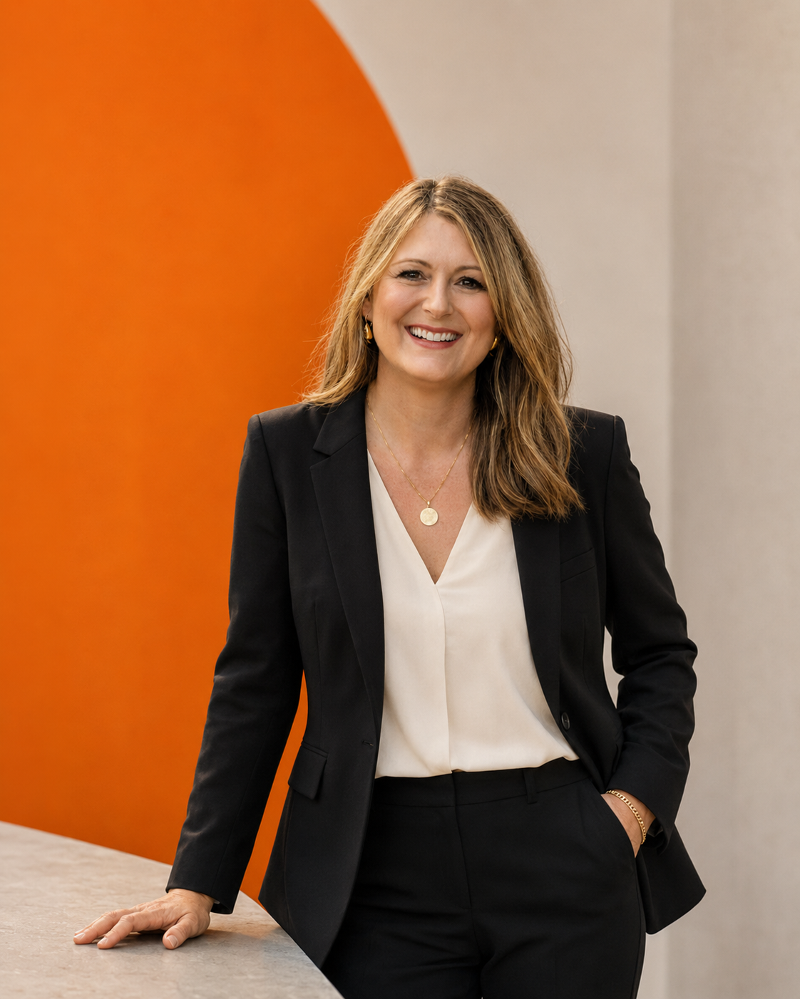 A woman in a black blazer and white blouse stands smiling with one hand on a counter and the other in her pocket, in front of an orange and gray background.