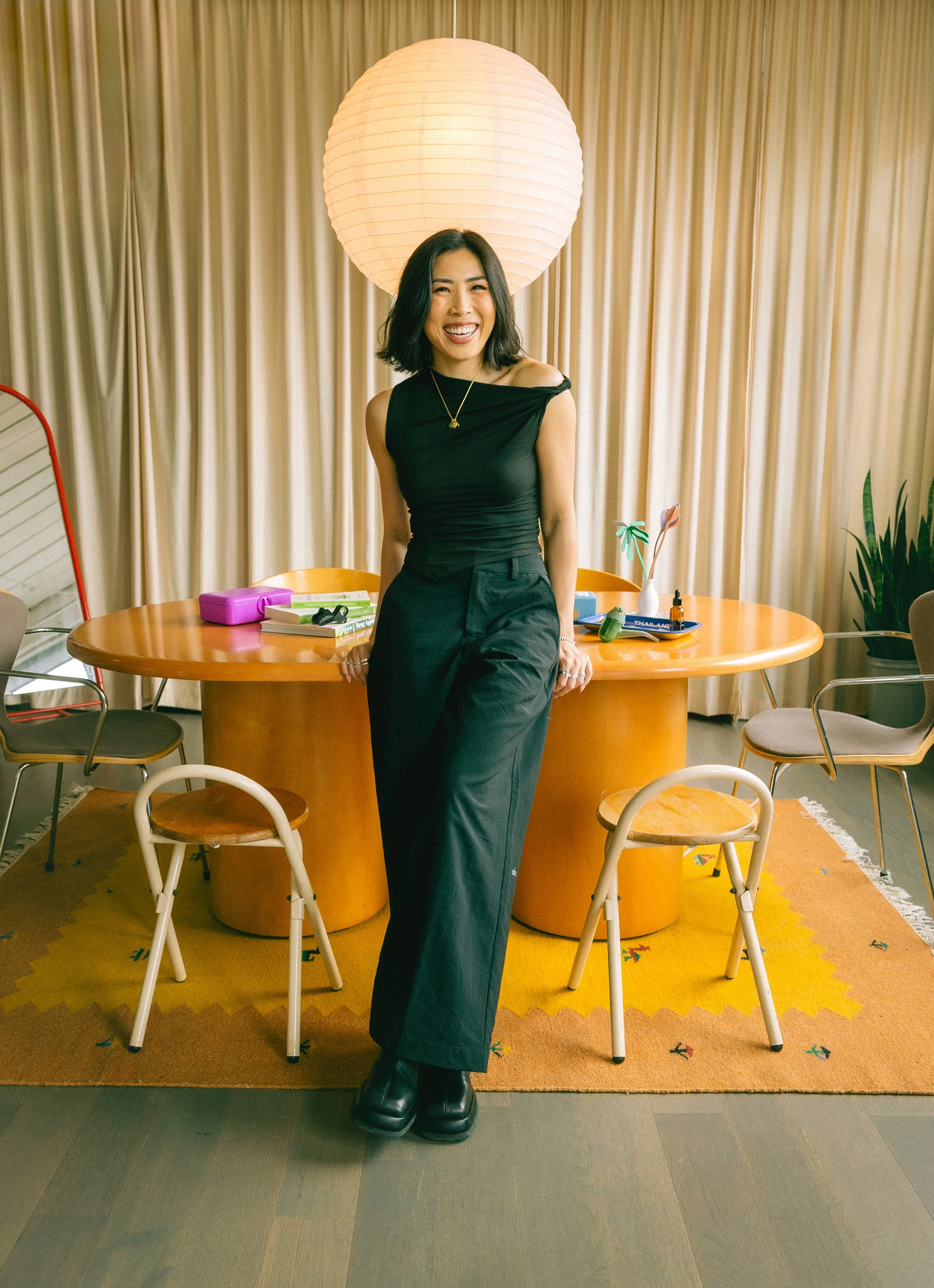 A woman with black hair smiling and standing indoors in front of a round wooden table with orange chairs. The background features beige curtains, a large paper lantern overhead, and a yellow rug with small colorful patterns.