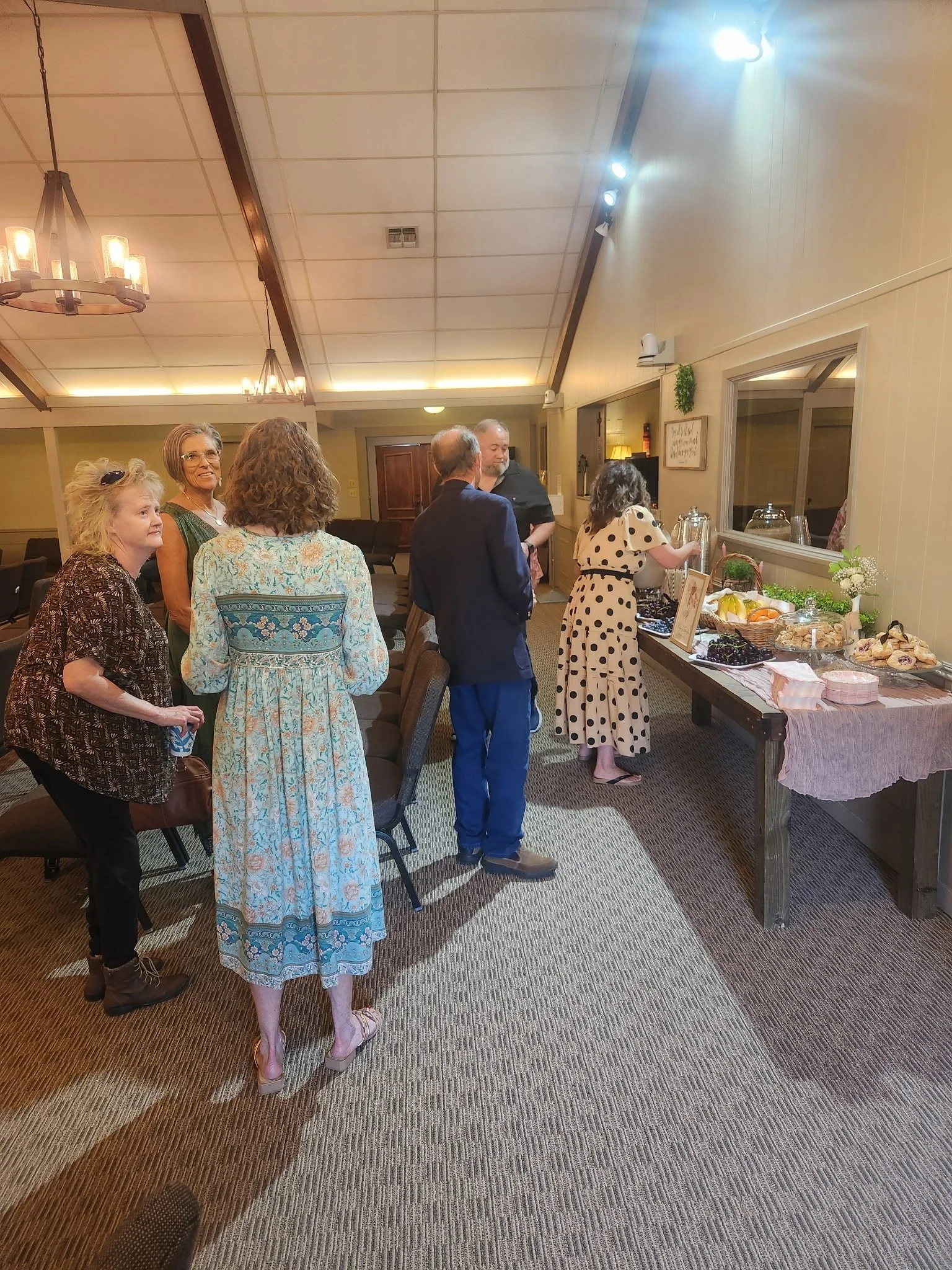 A group of people gathering around a table with food and drinks in a room with warm lighting and a wooden ceiling. There are six women and two men, some standing and some sitting, engaged in conversation.