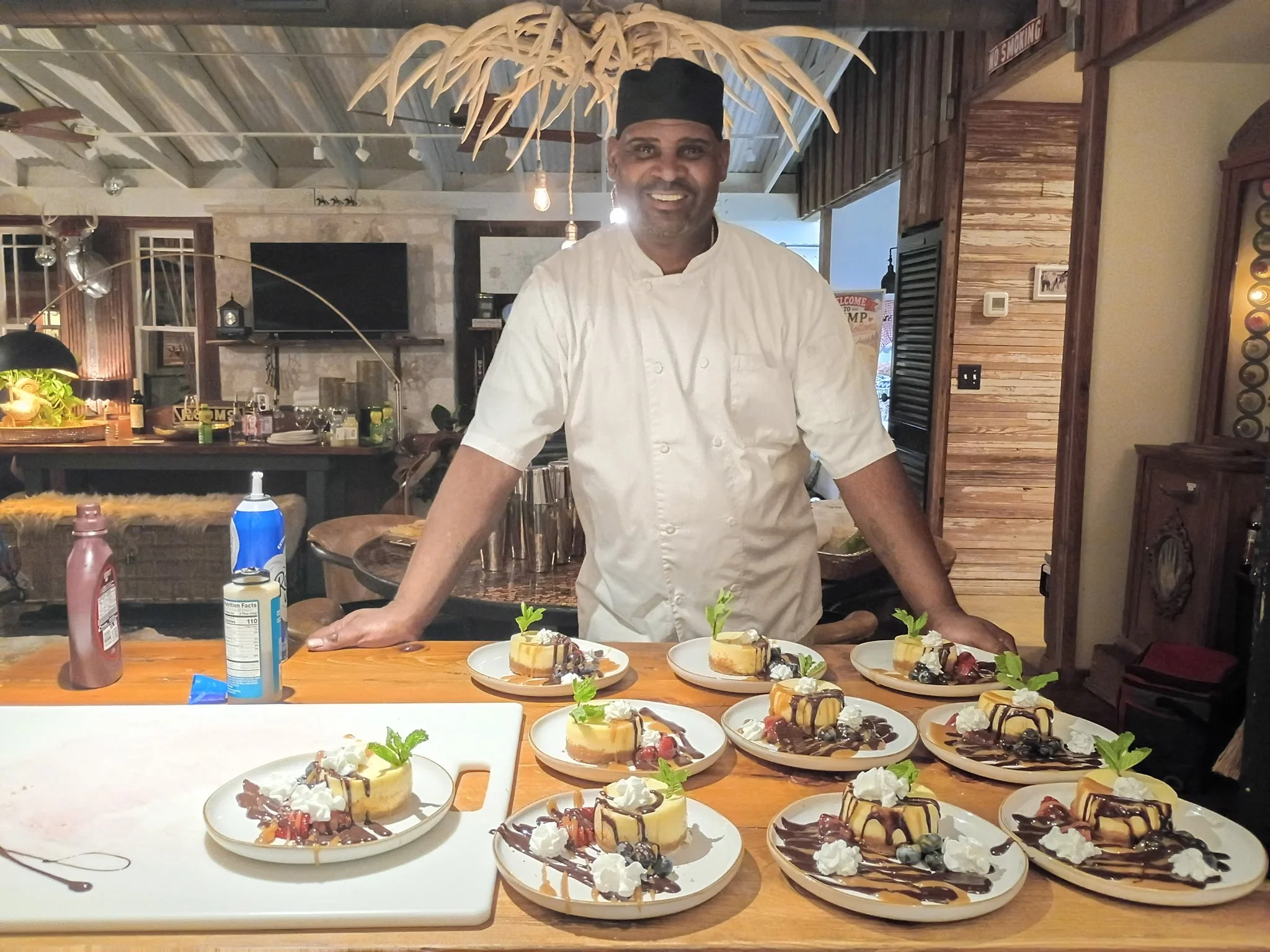 Chef Sheldon Jolley smiling behind a table of assorted desserts, including cheesecake topped with fruit, whipped cream, and chocolate drizzle, in a cozy restaurant setting.