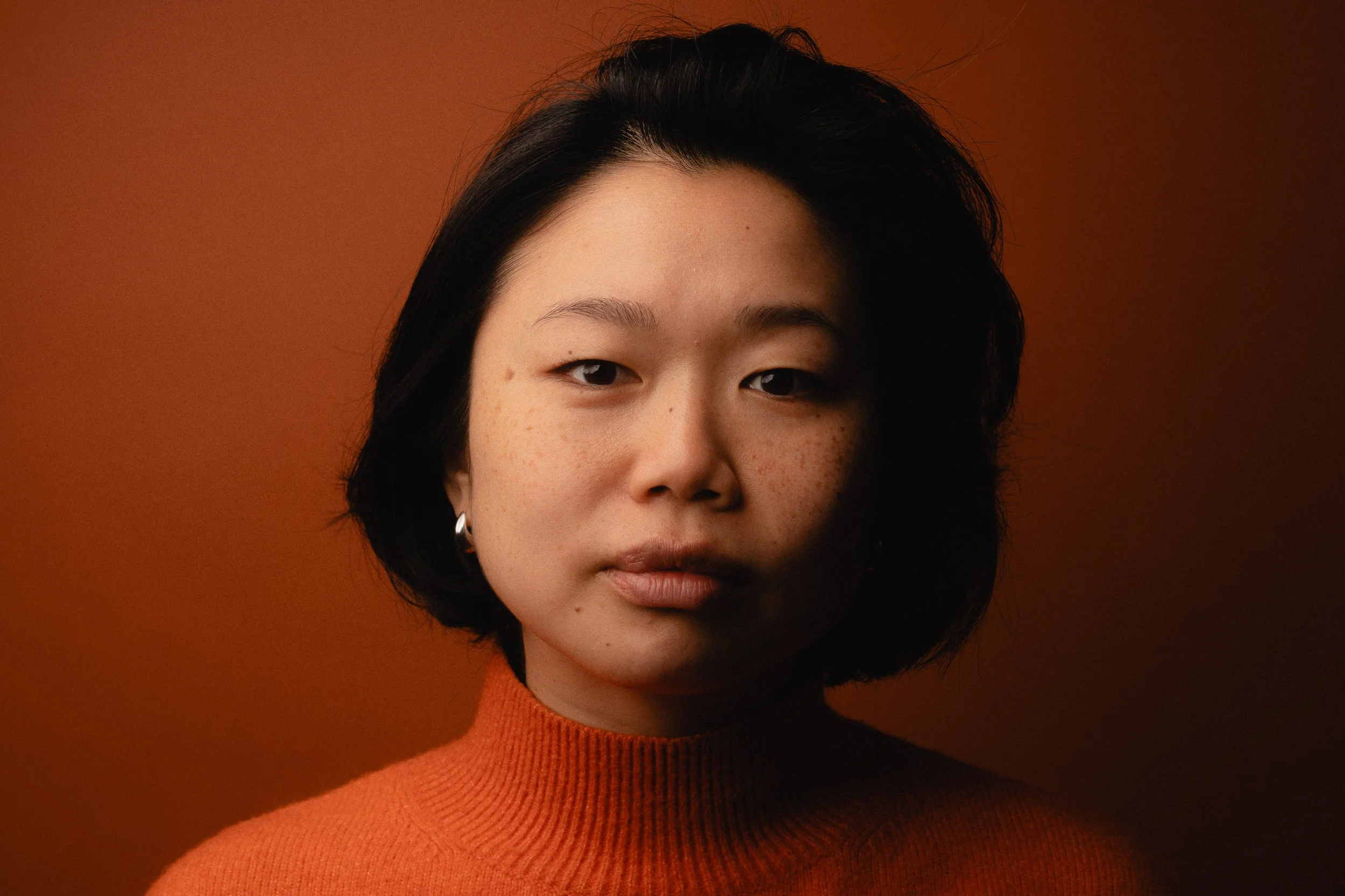 Portrait of a young woman with short black hair, wearing a burnt orange turtleneck sweater and silver hoop earrings against a dark orange background. Personal Branding Photography and Headshots in London.