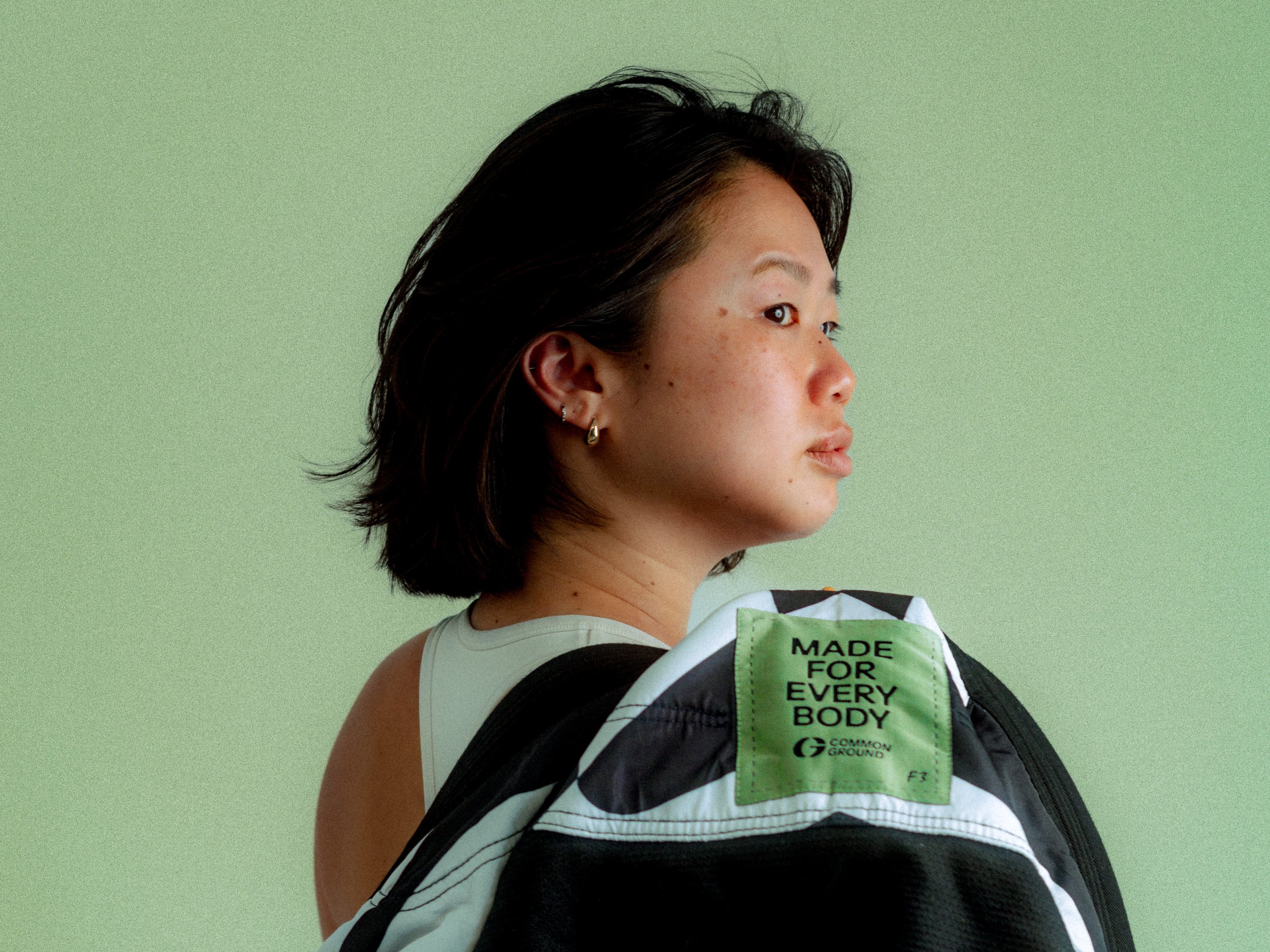 Young woman with dark hair and earrings looking to the right, carrying a jacket with a green tag against a green background. Personal Branding Photography and Headshots in London.