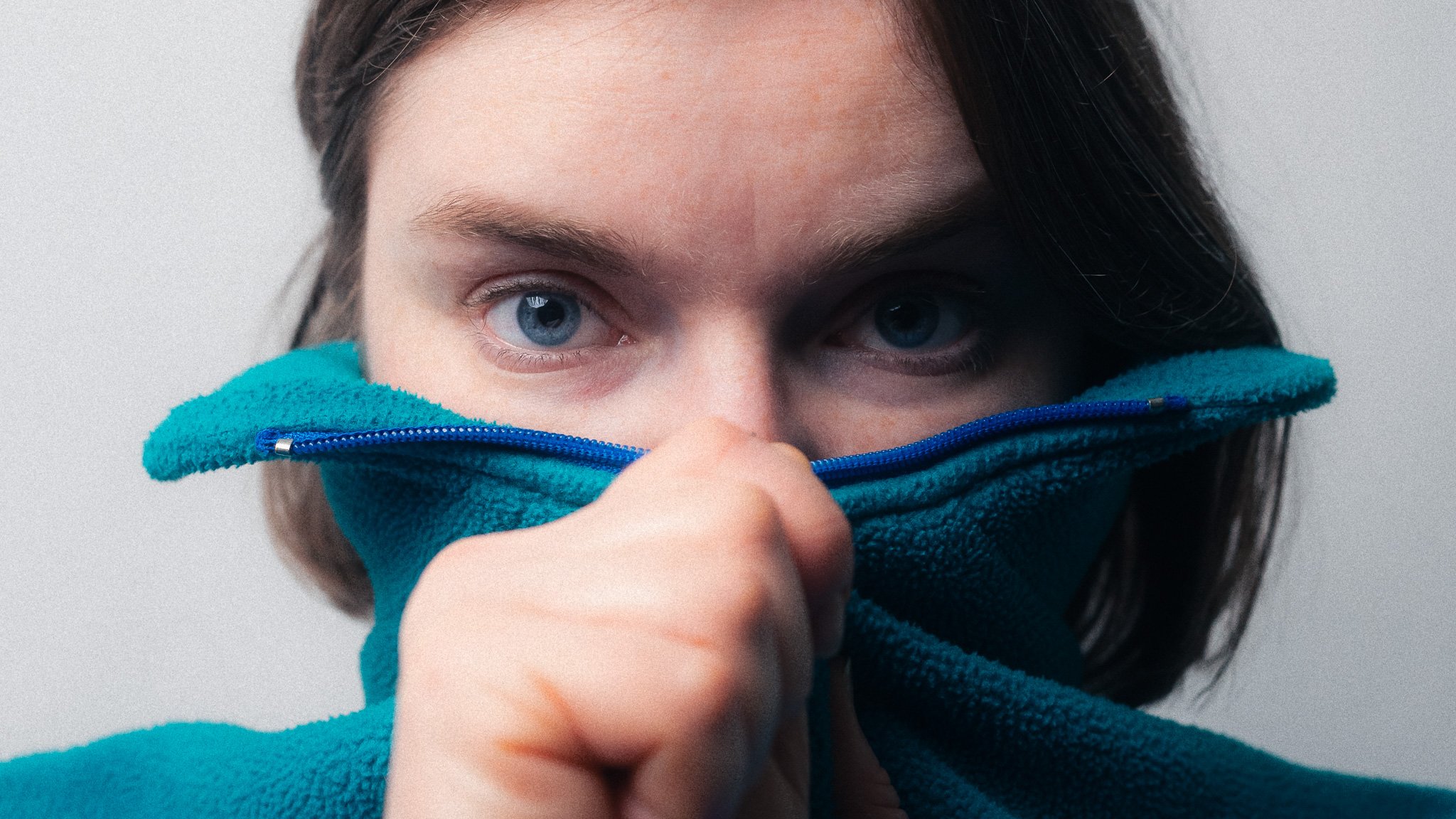 A woman with blue eyes and dark hair partially covered by a blue fleece jacket pulled up over her nose