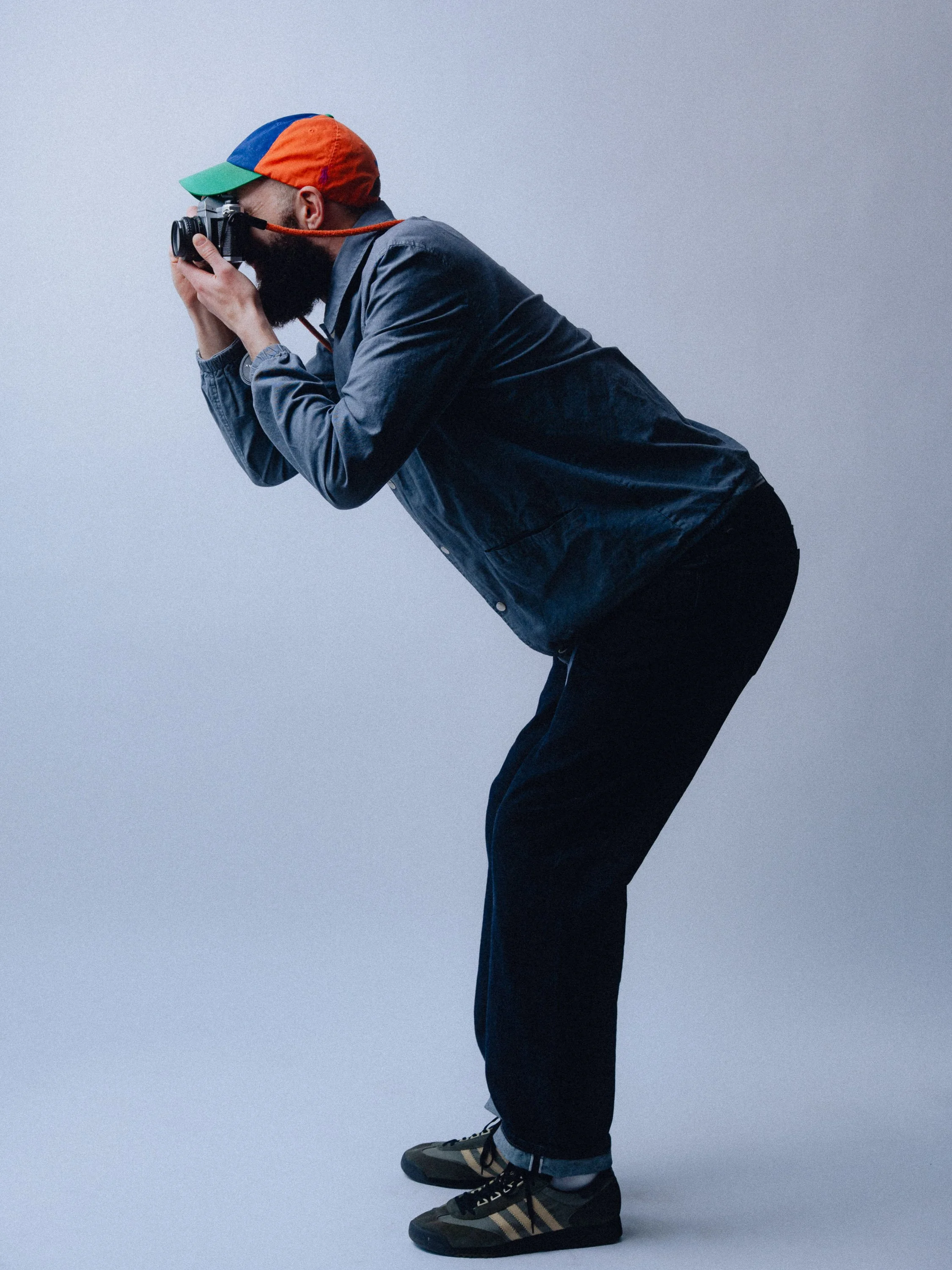 A man wearing a multicolored cap, black pants, a denim jacket, and sneakers, is crouched and taking a photograph with a camera against a plain light gray background. Personal Branding Photography and Headshots in London.