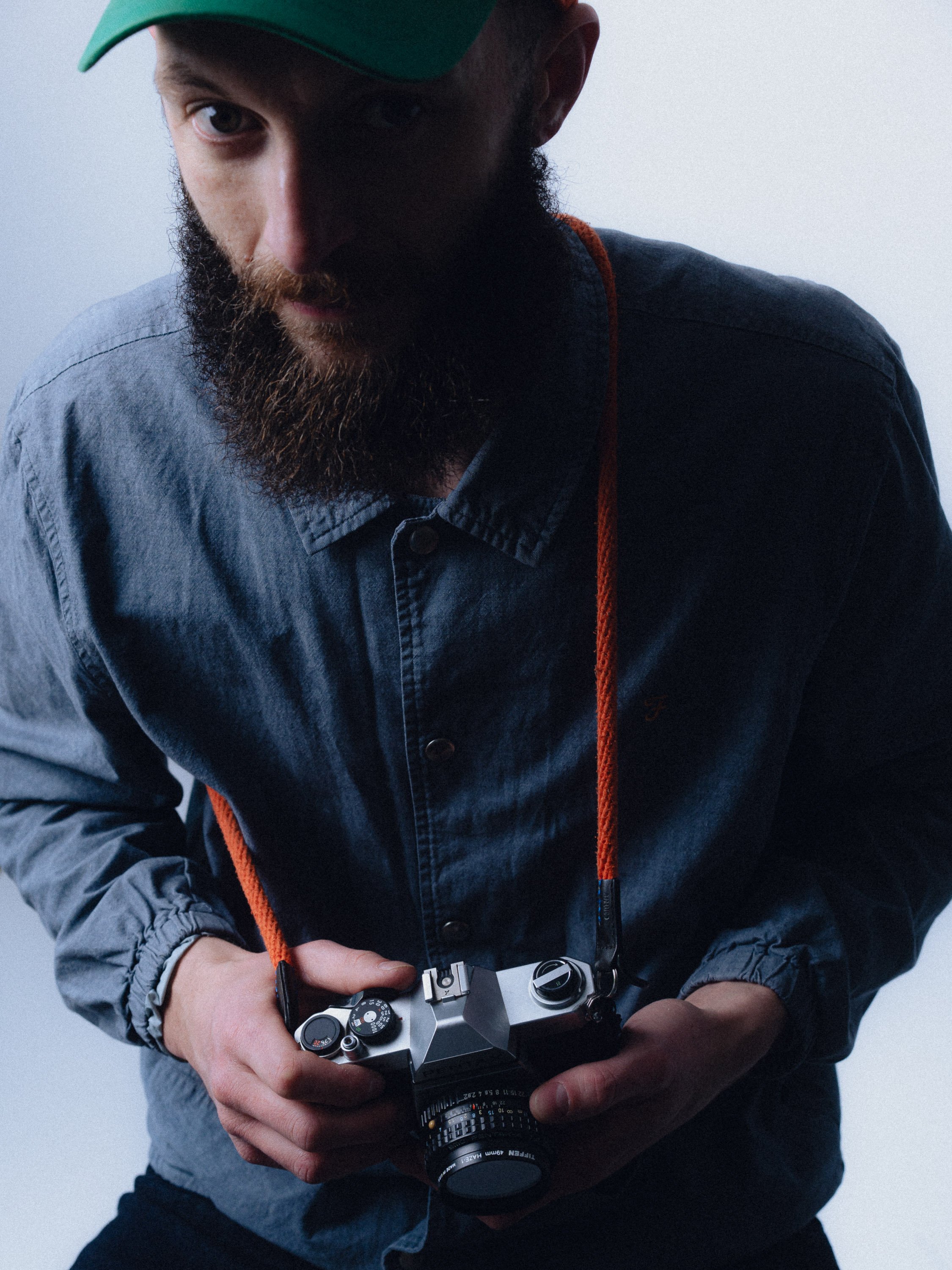 Man with a beard wearing a green cap and denim jacket holding a vintage camera with an orange strap. Personal Branding Photography and Headshots in London.