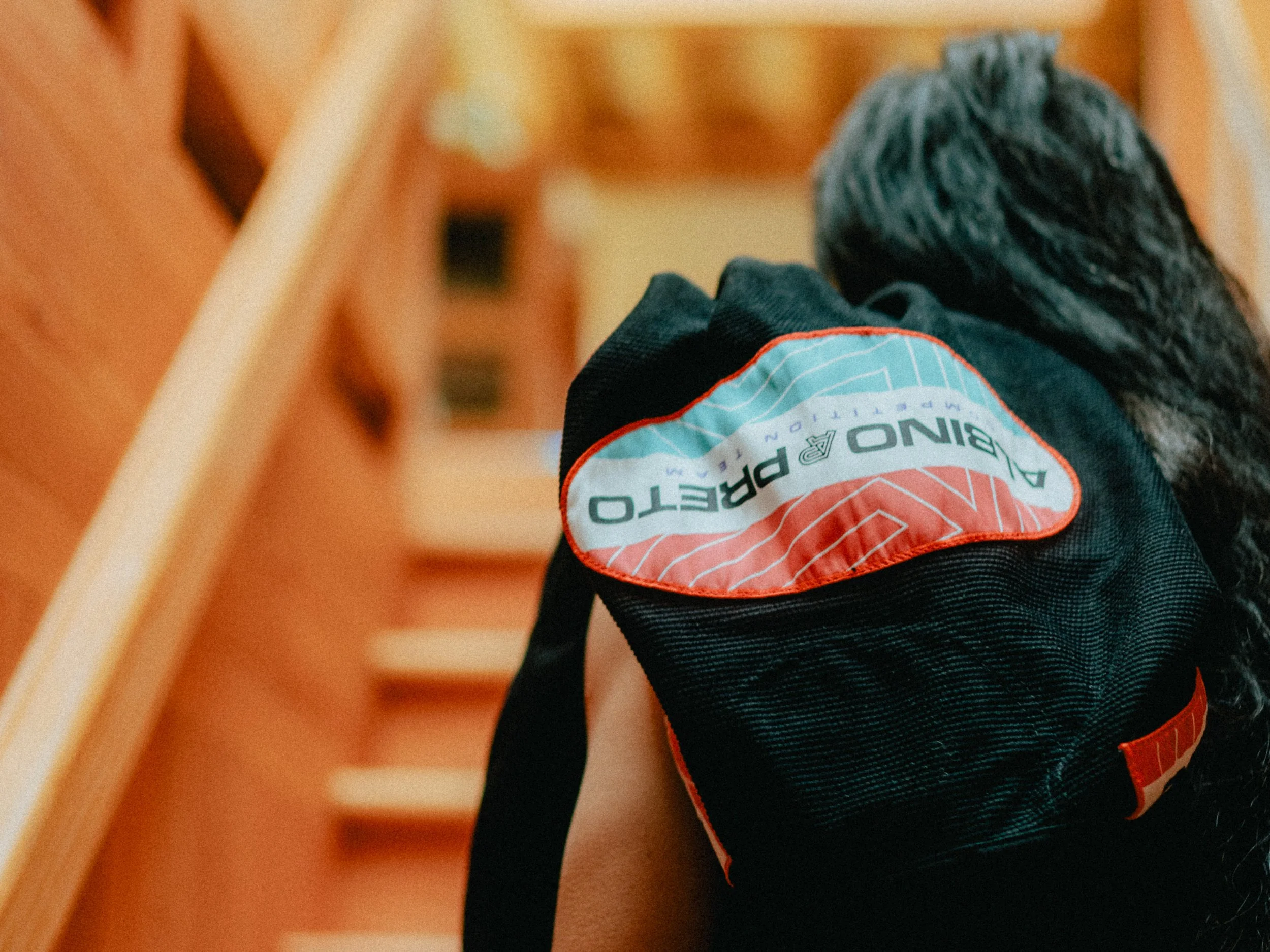 Close-up of a black backpack with a red and white patch labeled 'PING PHOTO' on the shoulder strap, with a blurred wooden staircase background. Personal Branding Photography and Headshots in London.