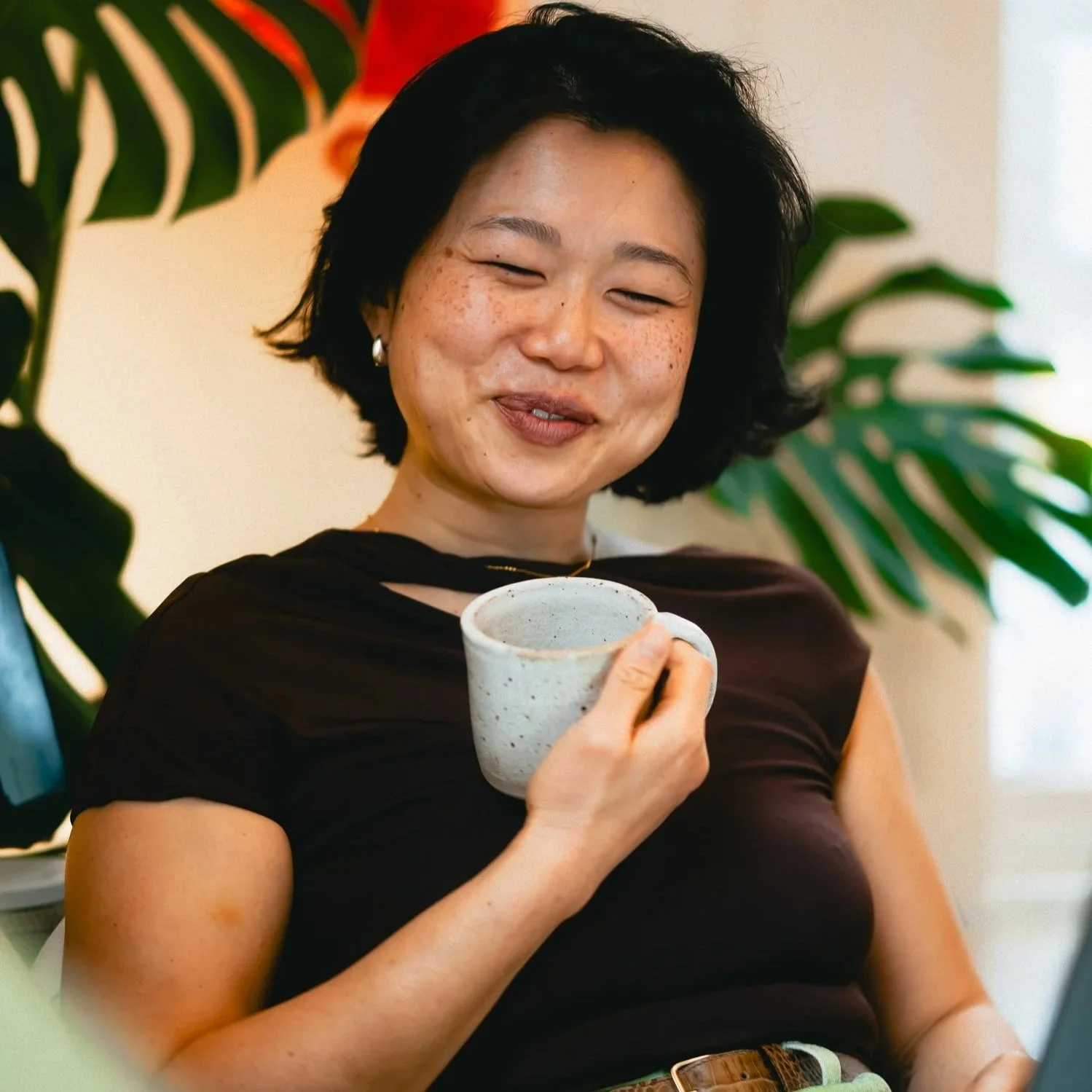 A woman with short black hair and freckles is smiling and holding a speckled ceramic mug, sitting indoors with large green plants in the background.