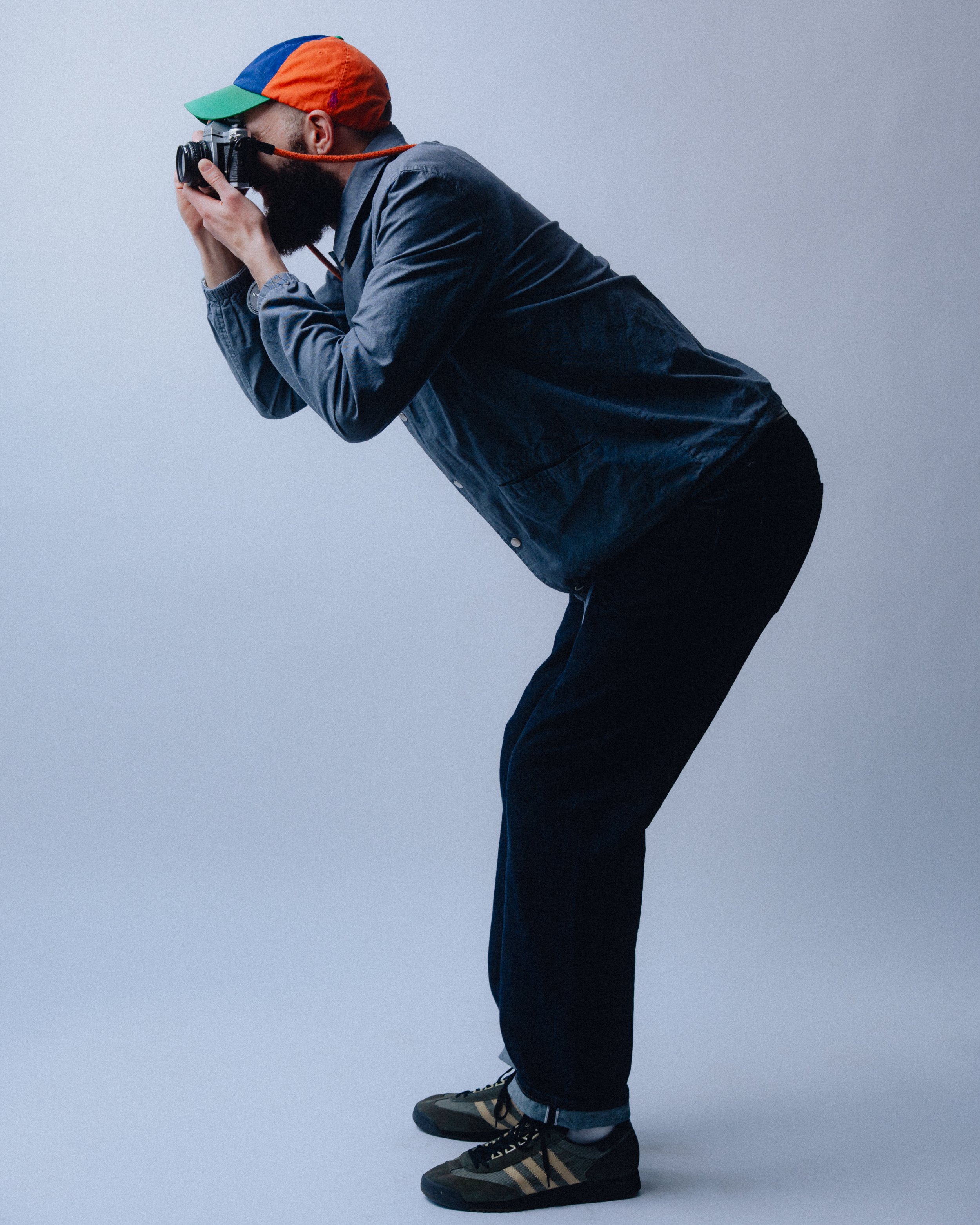 A man wearing a colorful cap, denim jacket, and black pants is leaning forward while taking a photograph with a camera.