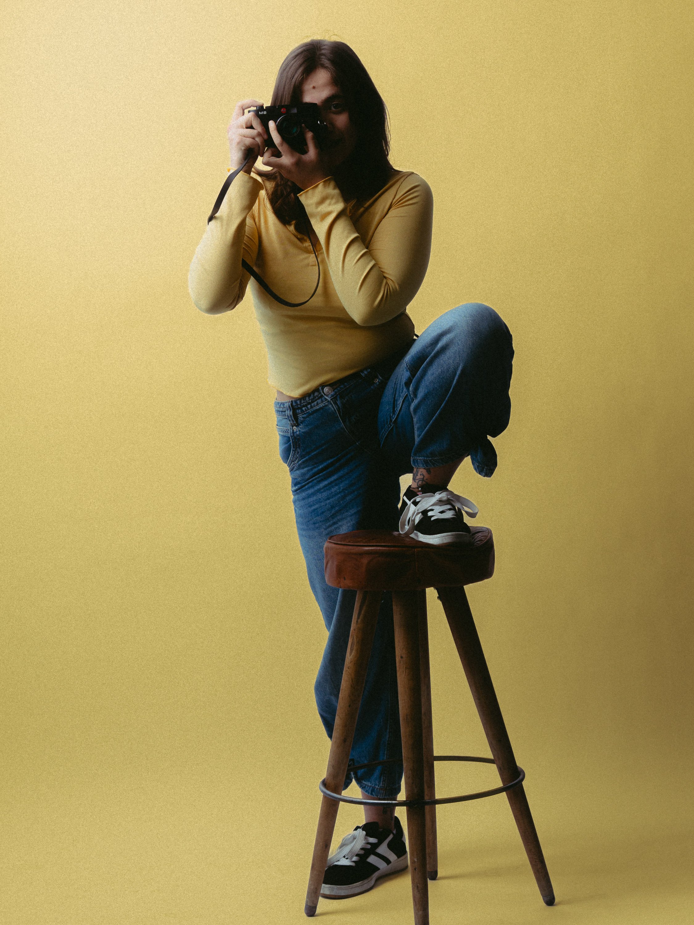 Woman with brown hair wearing a yellow long-sleeve shirt, blue jeans, and black and white sneakers, crouching with one foot on a wooden stool, taking a photo with a camera. Personal Branding Photography and Headshots in London.