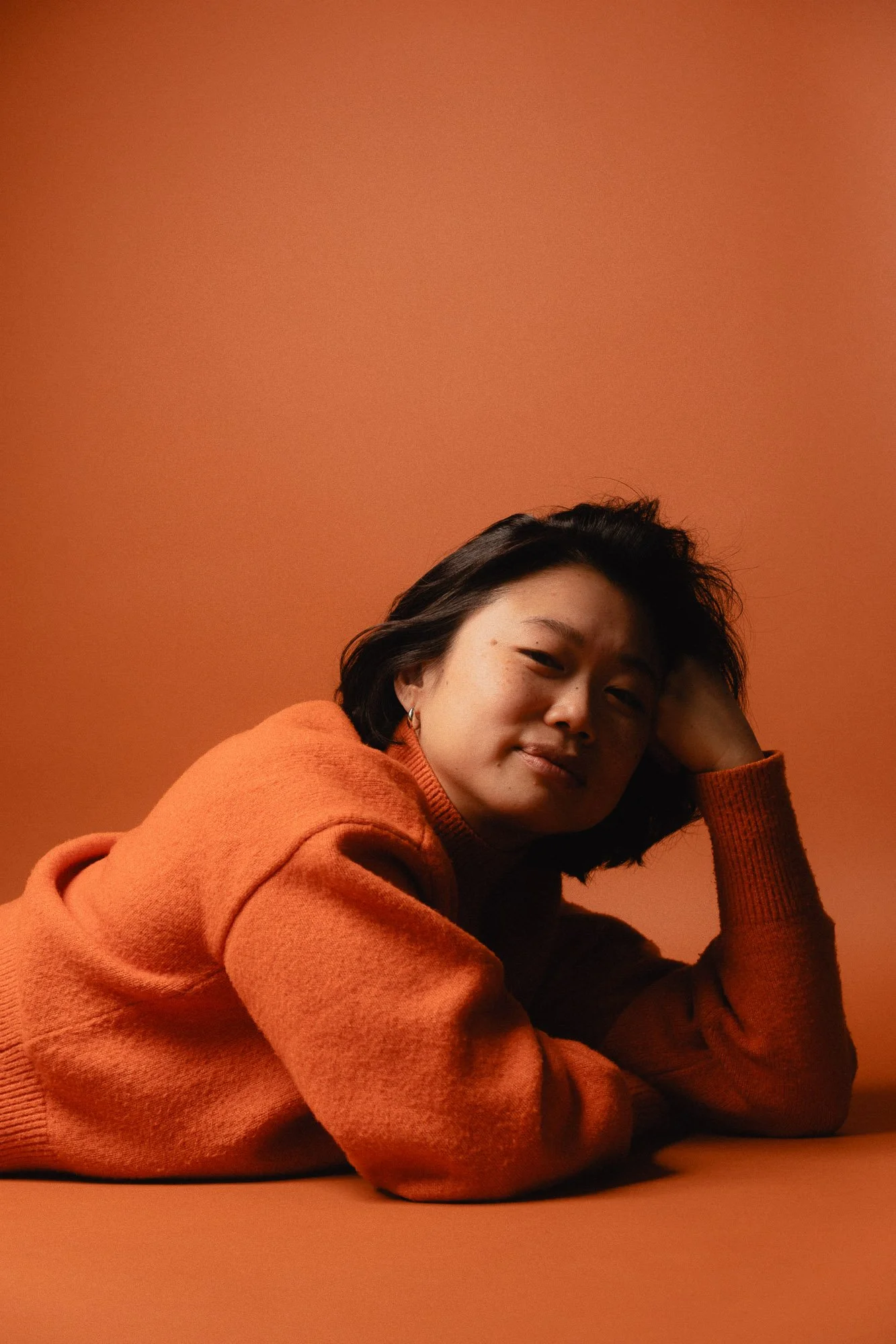 A woman with short dark hair wearing an orange sweater, resting her head on her hand, against an orange background.