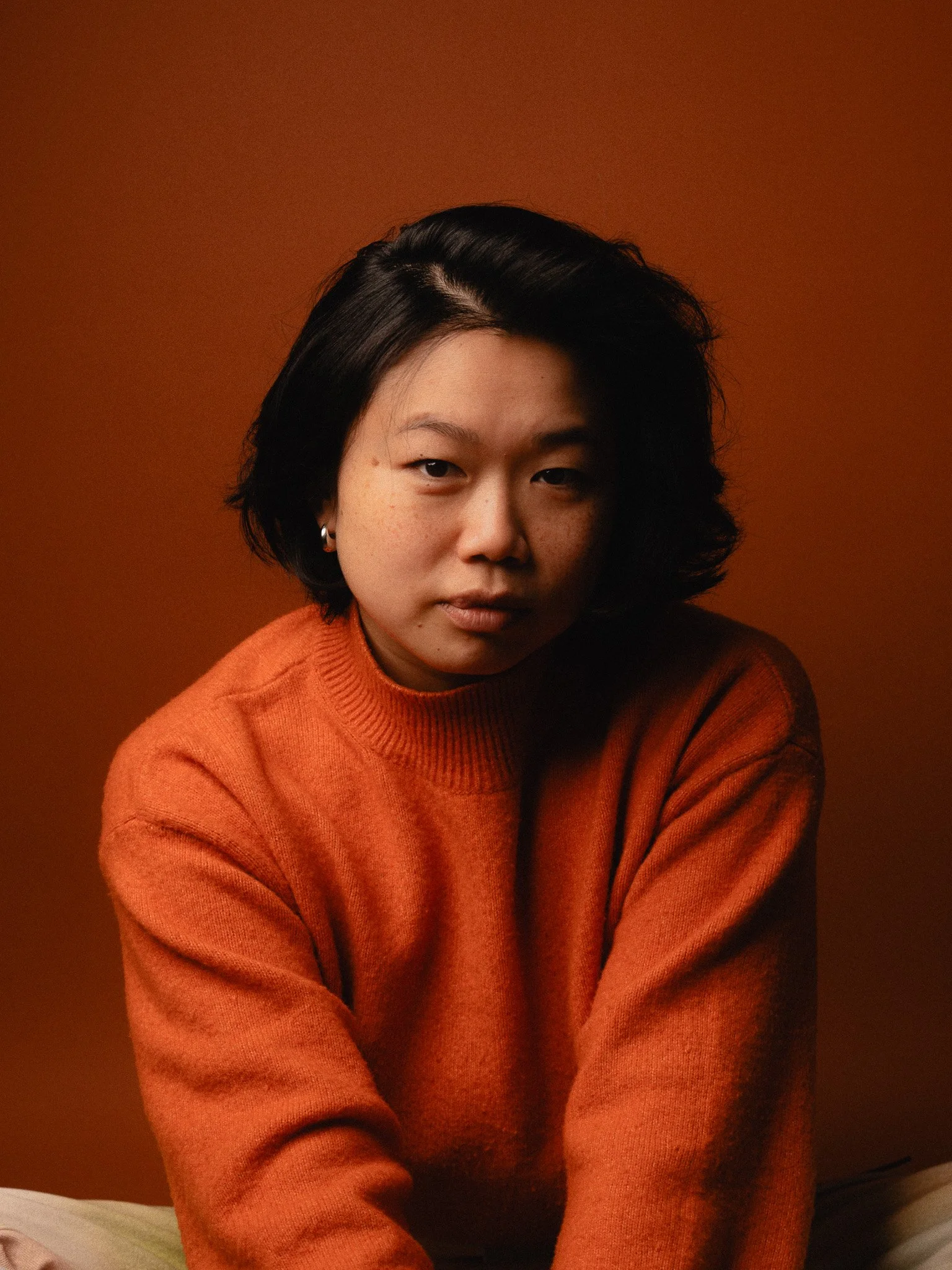 Portrait of a young woman with short black hair wearing an orange sweater against an orange background, looking directly at the camera.