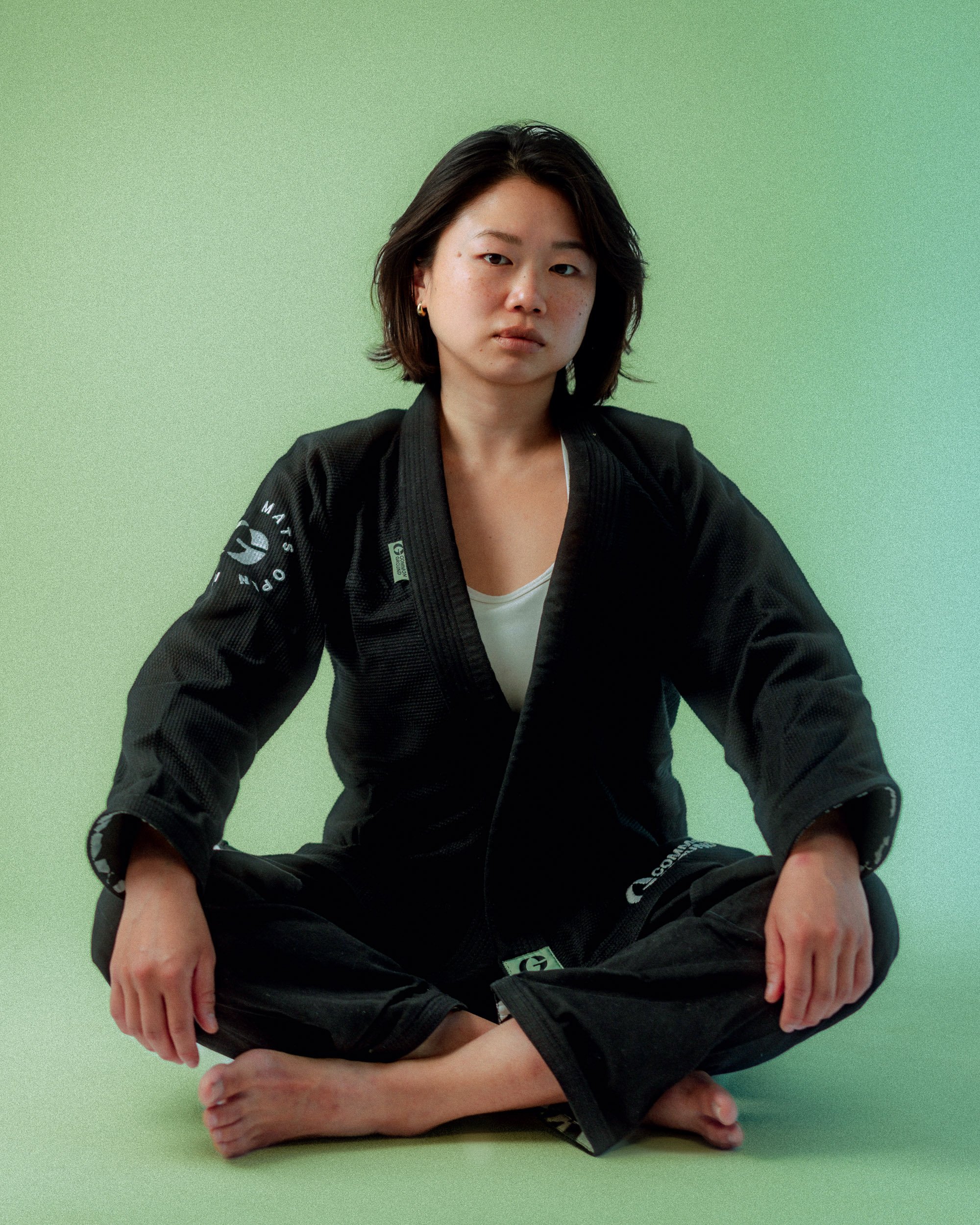 A woman sitting cross-legged on the floor wearing a black martial arts uniform against a green background. Personal Branding Photography and Headshots in London.