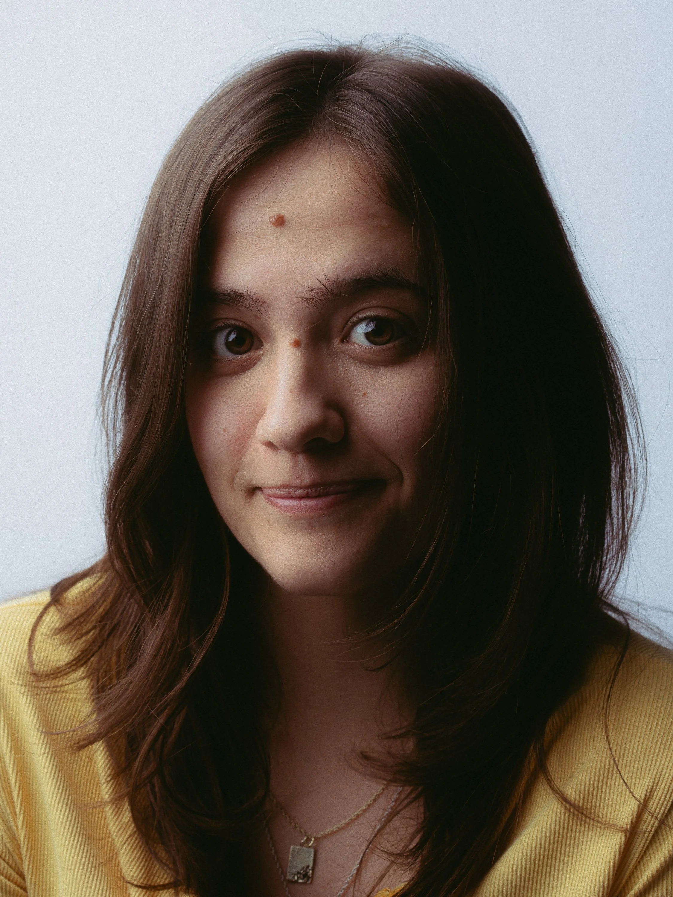 Close-up portrait of a young woman with brown hair, wearing a yellow top and a gold necklace, smiling slightly against a plain light background. Personal Branding Photography and Headshots in London.