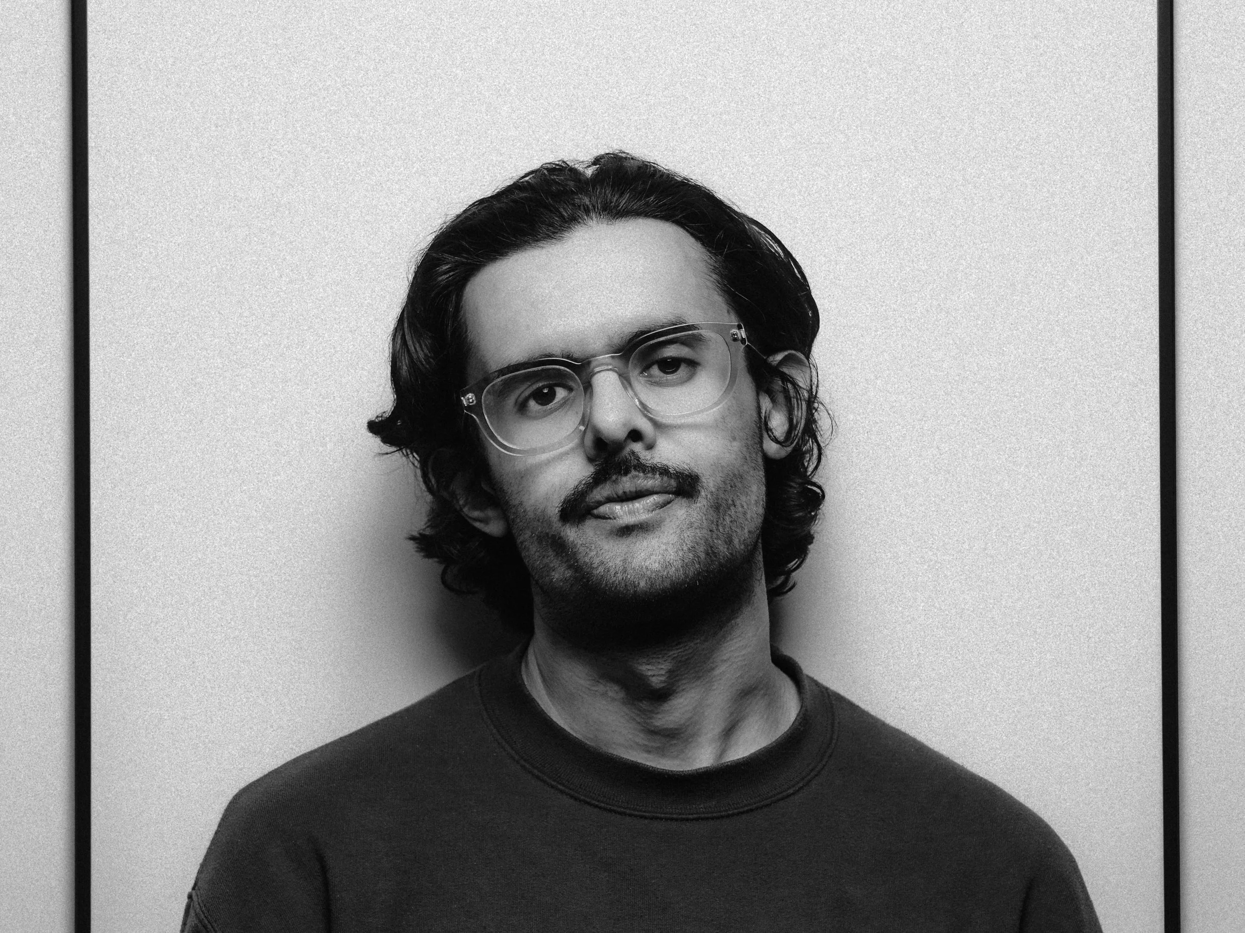 Black and white portrait of a man with wavy hair, glasses, and facial hair wearing a dark shirt, standing against a plain light background. Personal Branding Photography and Headshots in London.
