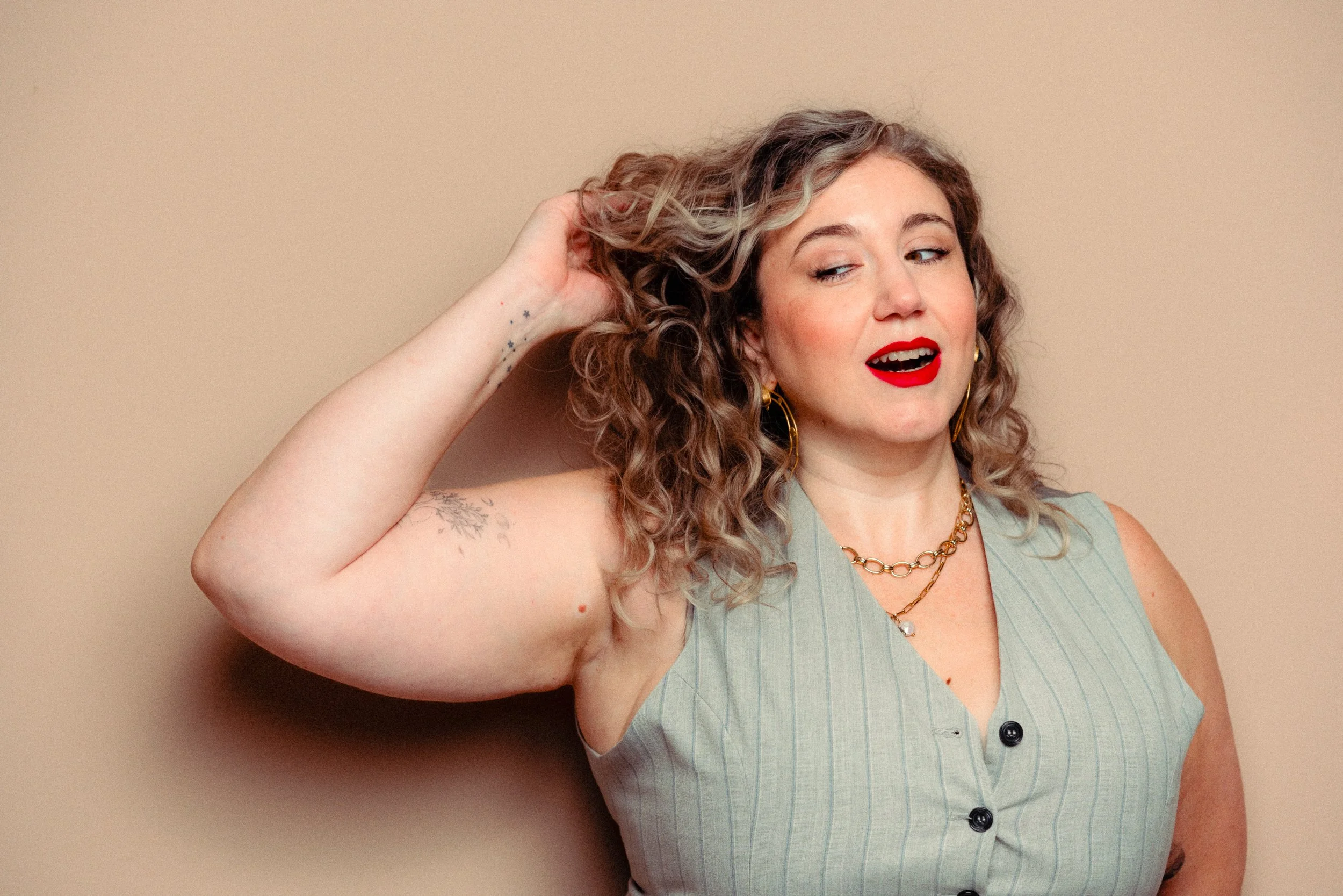 A woman with curly hair and light skin wearing a sleeveless gray vest with black buttons, red lipstick, gold hoop earrings, and layered gold necklaces. Personal Branding Photography and Headshots in London.