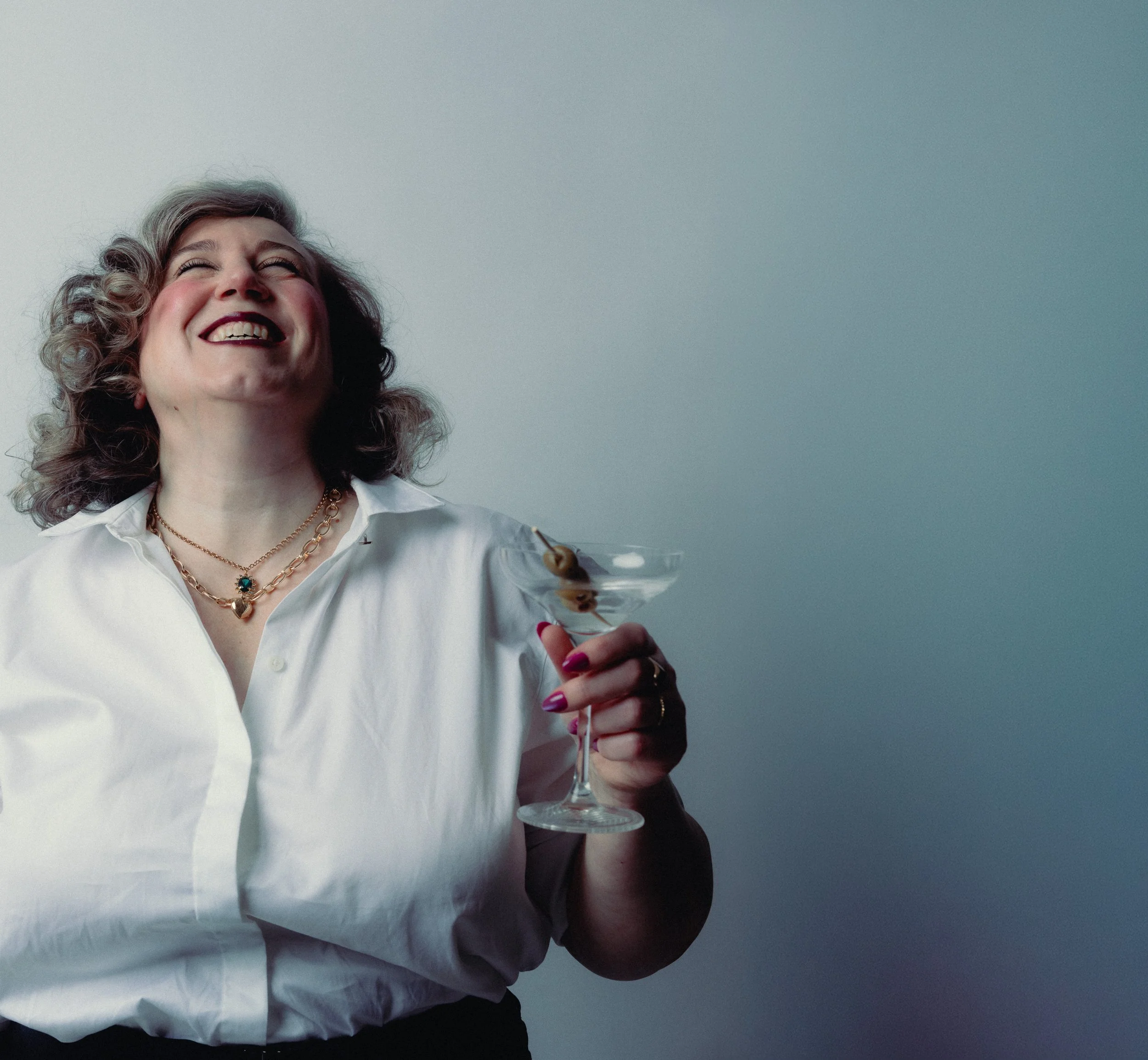 A woman with curly hair and layered necklaces, wearing a white button-up shirt, smiling and holding a martini glass with an olive garnish, against a plain gray background. Personal Branding Photography and Headshots in London.