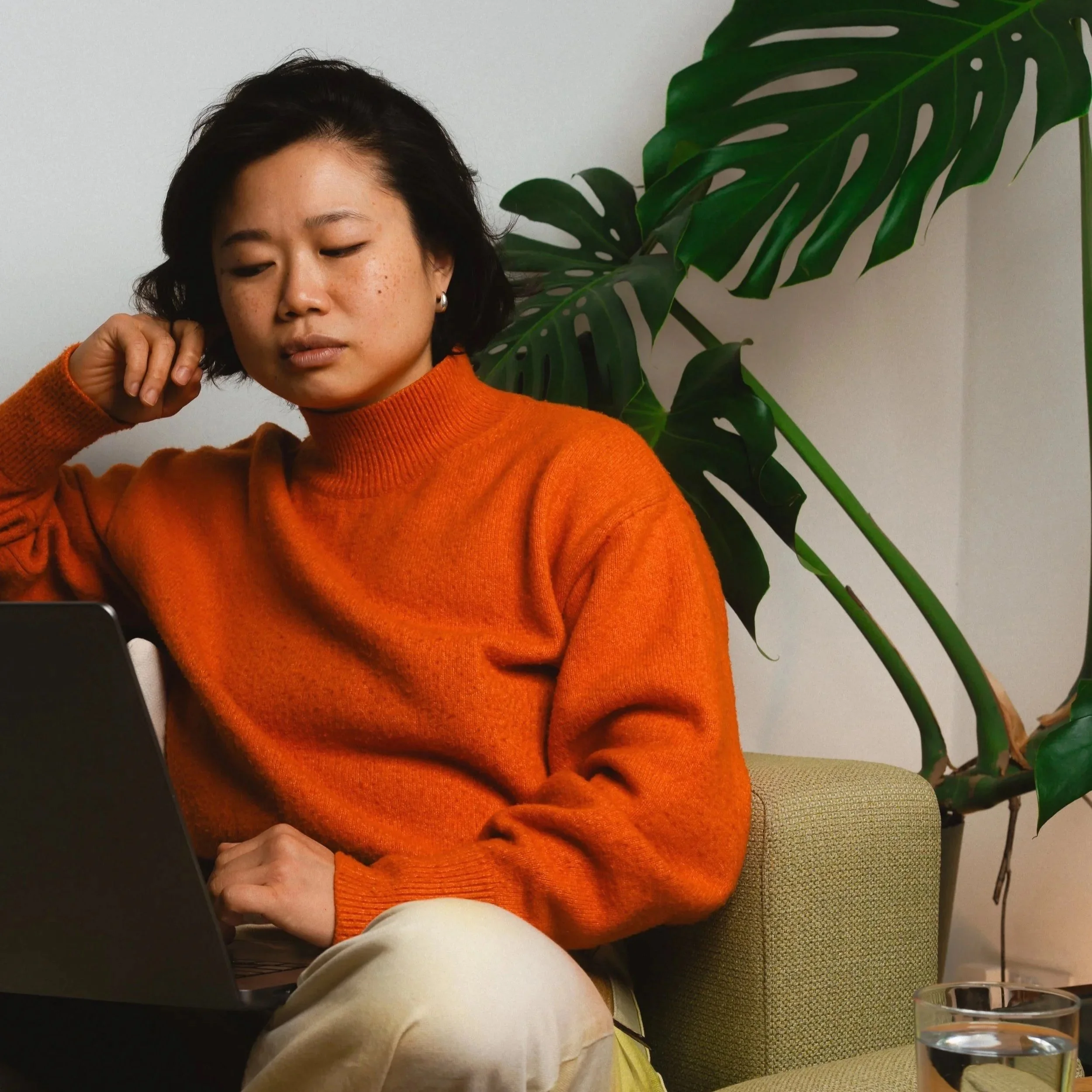 A woman with short black hair, wearing an orange sweater, sitting on a green armchair, working on a laptop, with a large green monstera plant behind her.