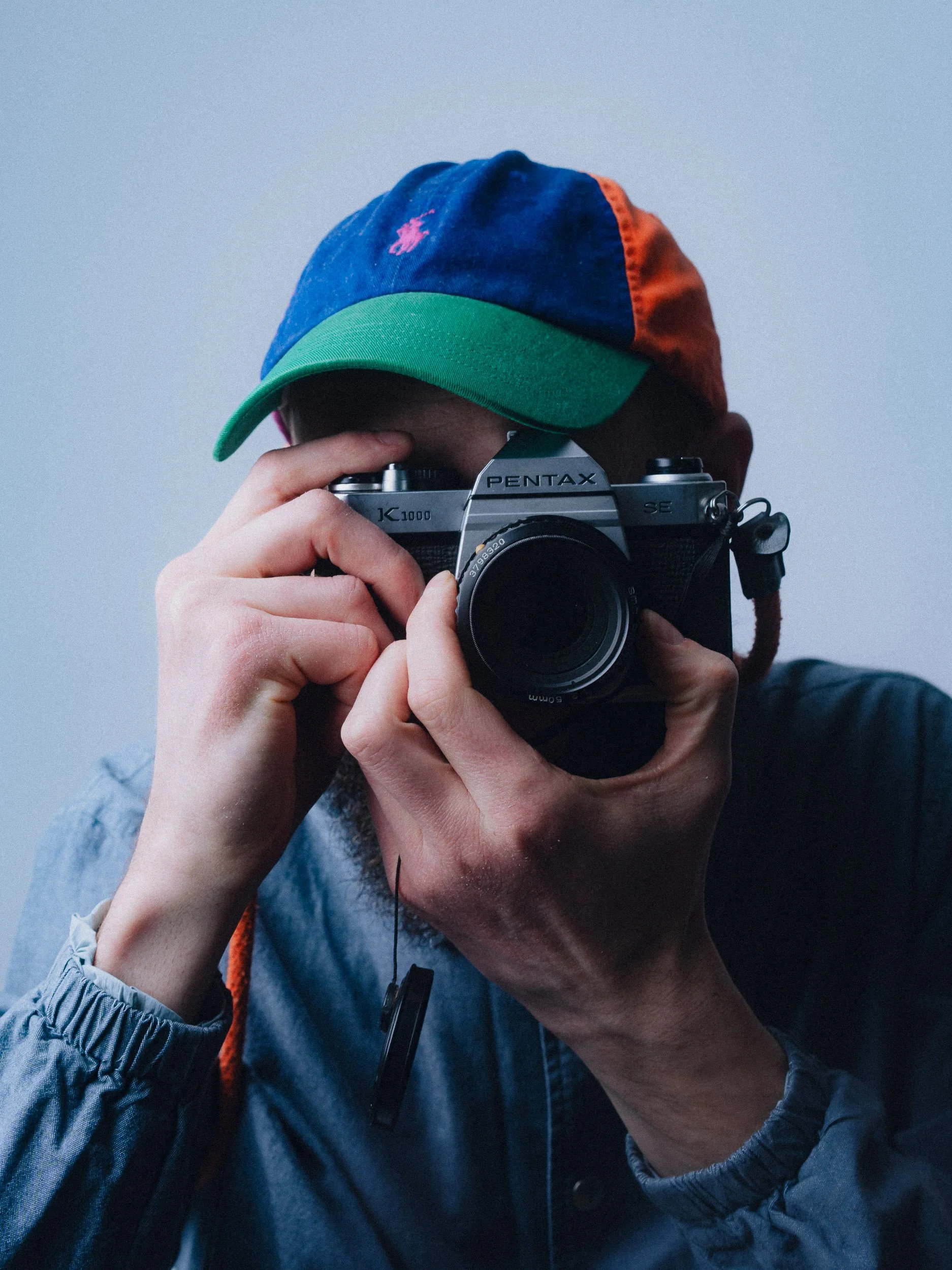 Person wearing a colorful cap and a gray jacket, taking a photo with a vintage Pentax film camera. Personal Branding Photography and Headshots in London.