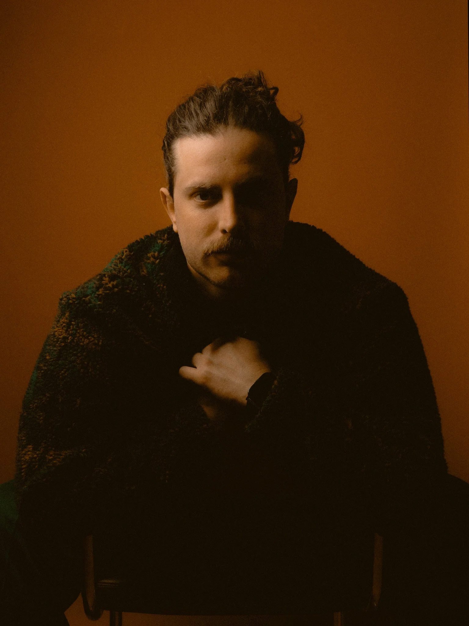 Portrait of a man with curly hair and mustache, sitting against a warm brown background, wearing a textured dark sweater, with one hand on his chest and looking directly at the camera.