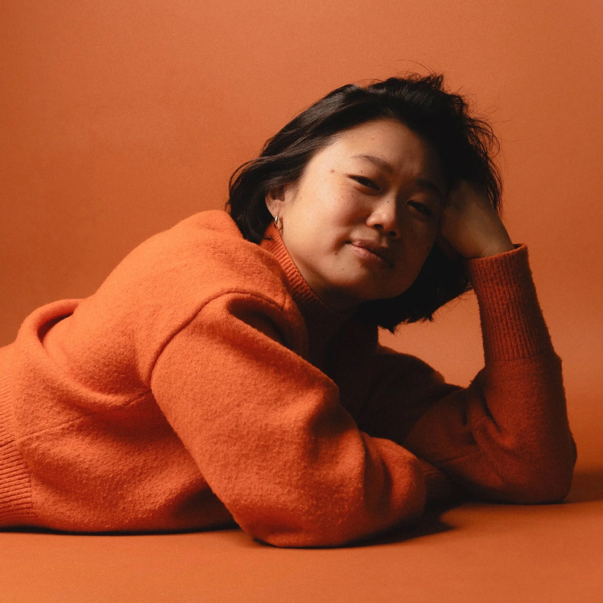 A woman with short dark hair wearing an orange sweater, resting her head on her hand, against an orange background.