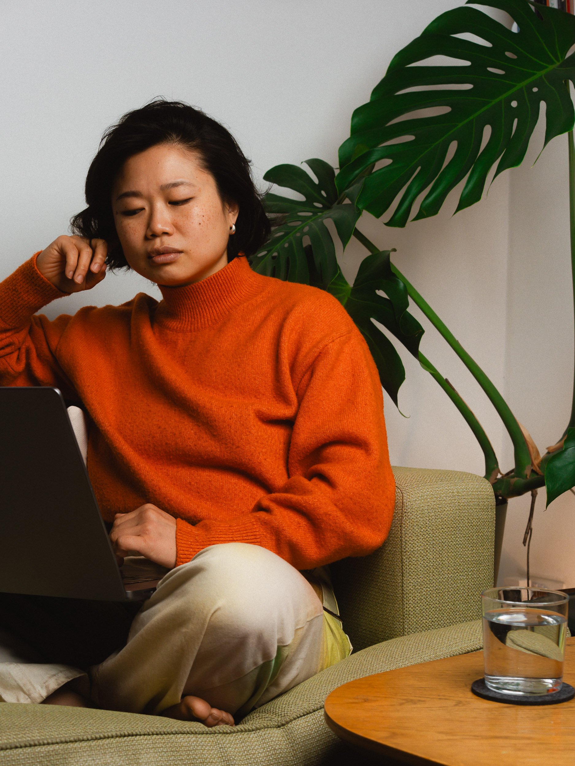 A woman wearing an orange sweater and beige pants sitting on a green couch, working on a laptop, with a large Monstera plant behind her and a glass of water on a wooden table beside her. Personal Branding Photography and Headshots in London.