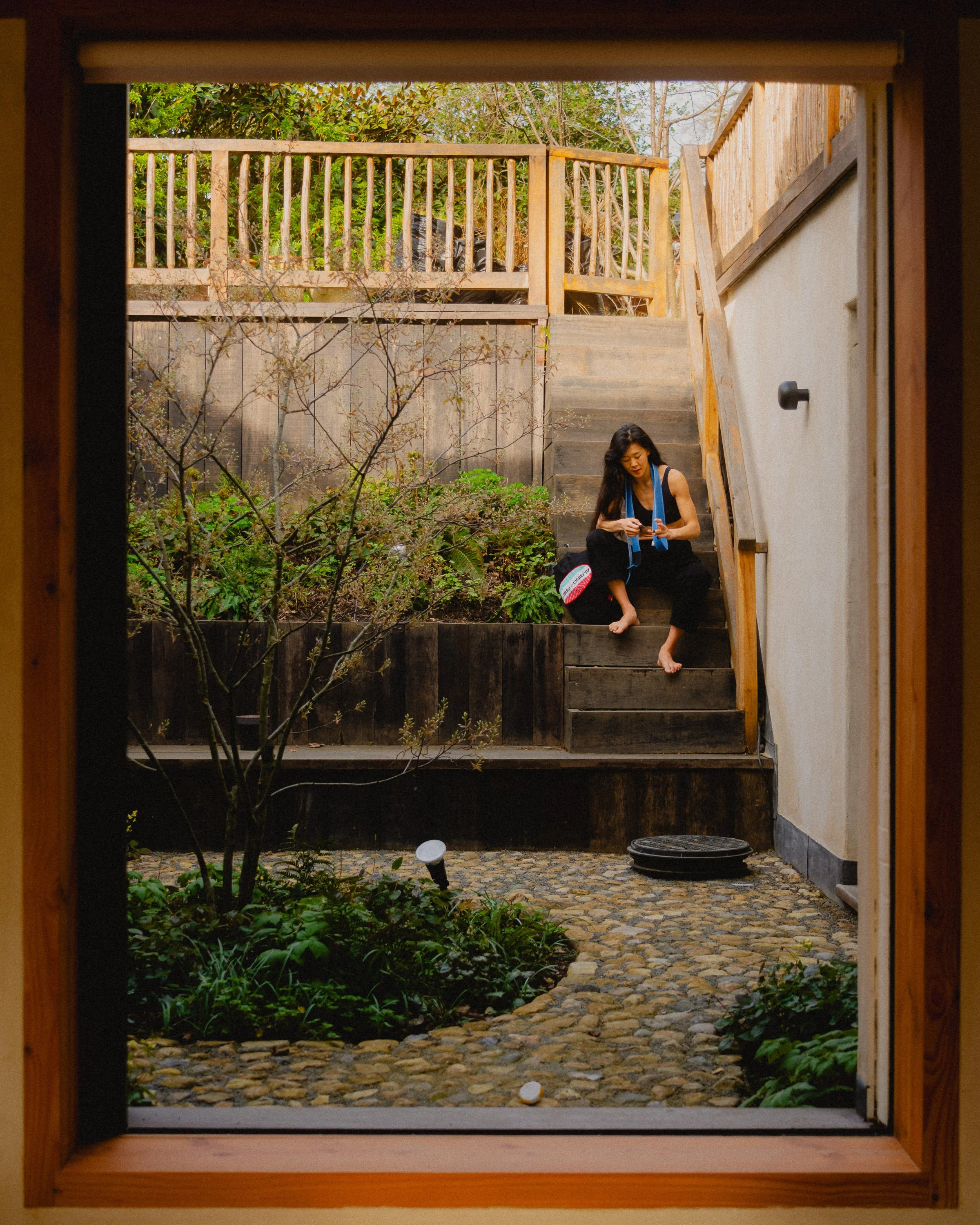 A woman sitting on outdoor stairs in a backyard, looking at her phone. The yard has a small garden and a stone pathway, framed by a wooden deck and railing. Personal Branding Photography and Headshots in London.