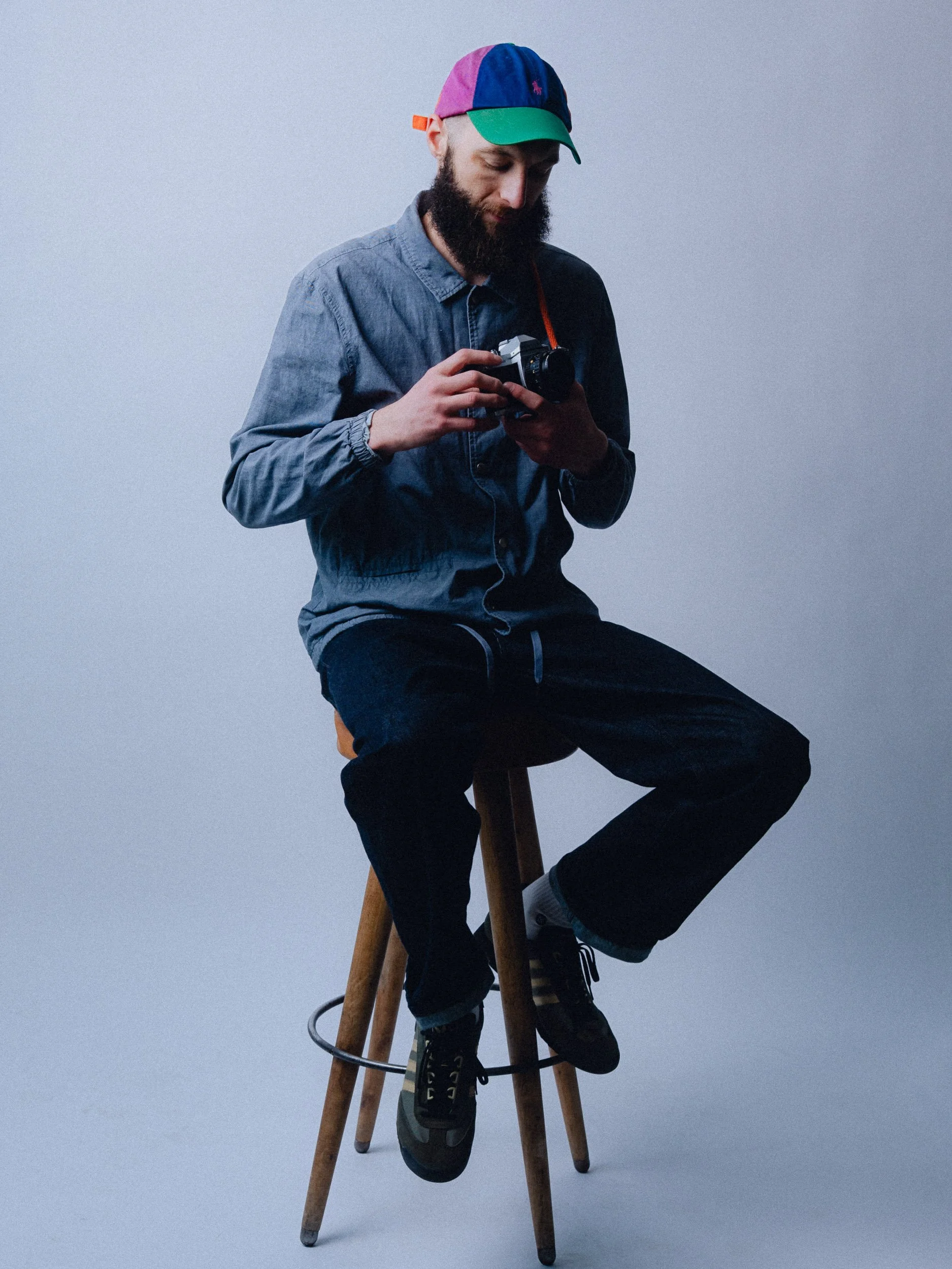 A man with a beard sitting on a wooden stool, looking at a camera, wearing a colorful cap, a denim shirt, black pants, and sneakers against a plain background. Personal Branding Photography and Headshots in London.
