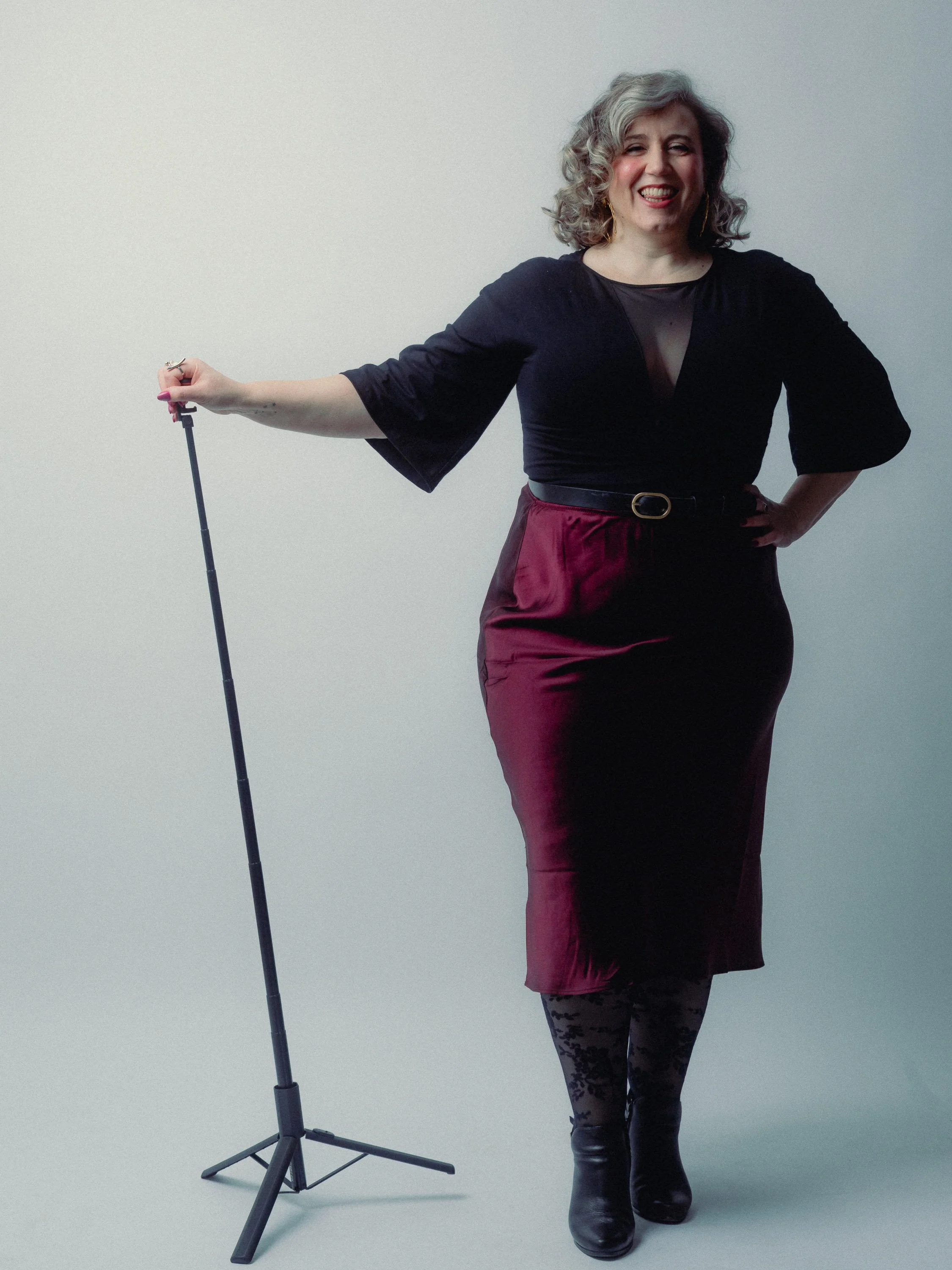 A woman with curly gray hair standing against a plain light background. She is smiling, holding a microphone stand in one hand. Personal Branding Photography and Headshots in London.
