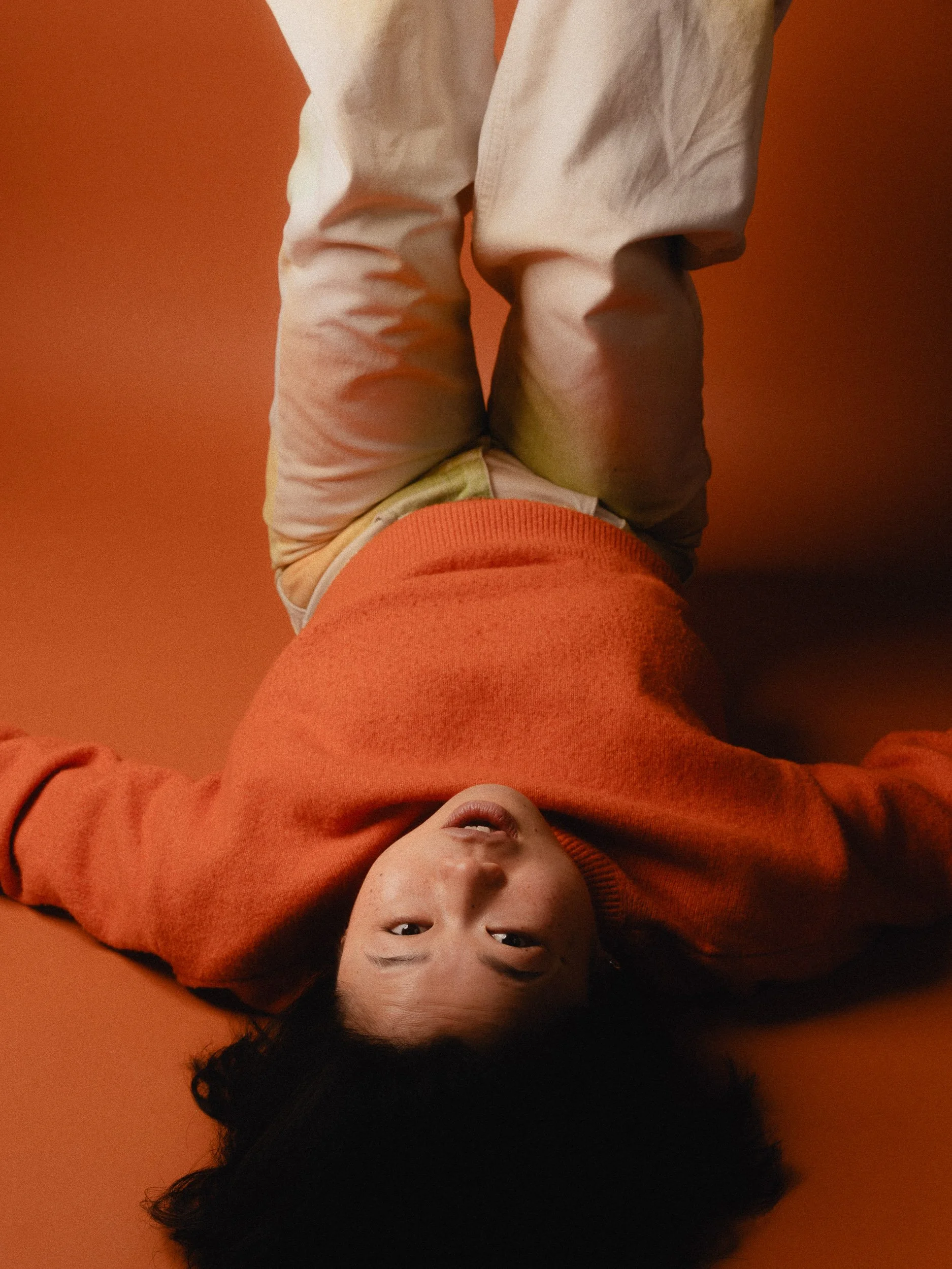 A young boy lying on his back on an orange surface, wearing an orange sweater and beige pants, looking up at the camera with a neutral expression. Personal Branding Photography and Headshots in London.