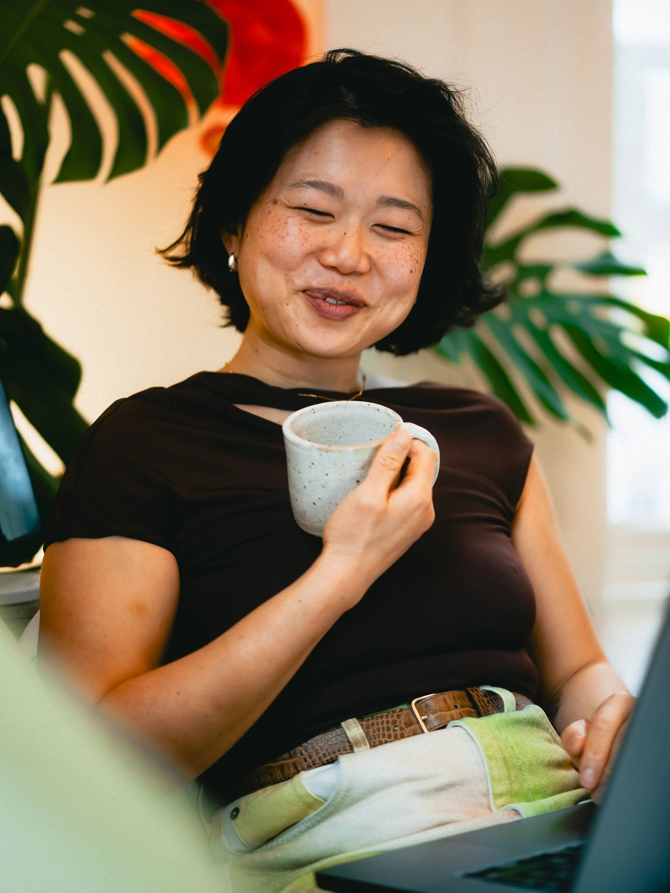 A woman with short black hair and freckles, smiling and holding a ceramic mug, sitting near a laptop with large green leaves in the background. Personal Branding Photography and Headshots in London.