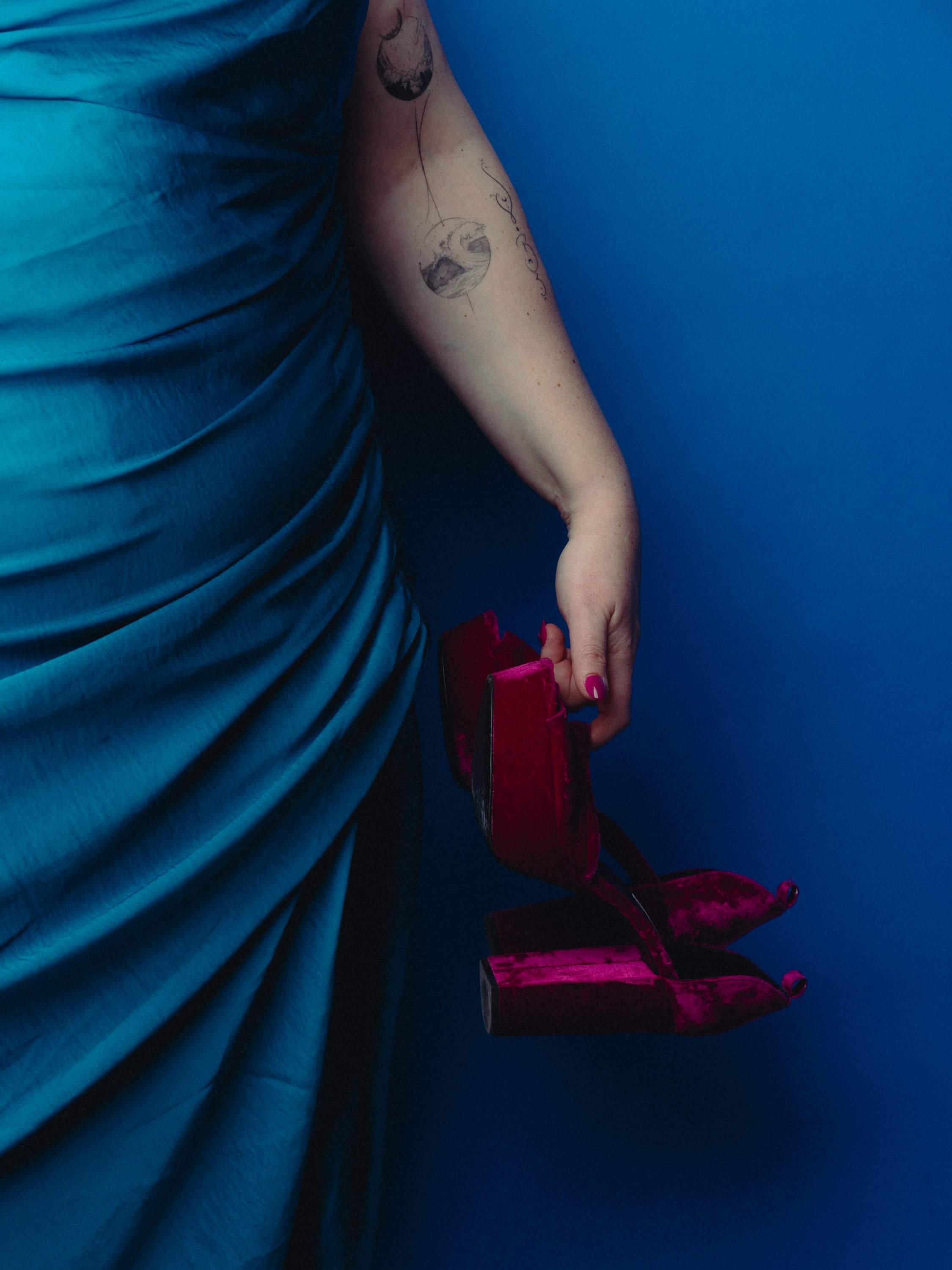 A person wearing a blue dress holding a small red velvet high heel shoe against a blue background. Personal Branding Photography and Headshots in London.