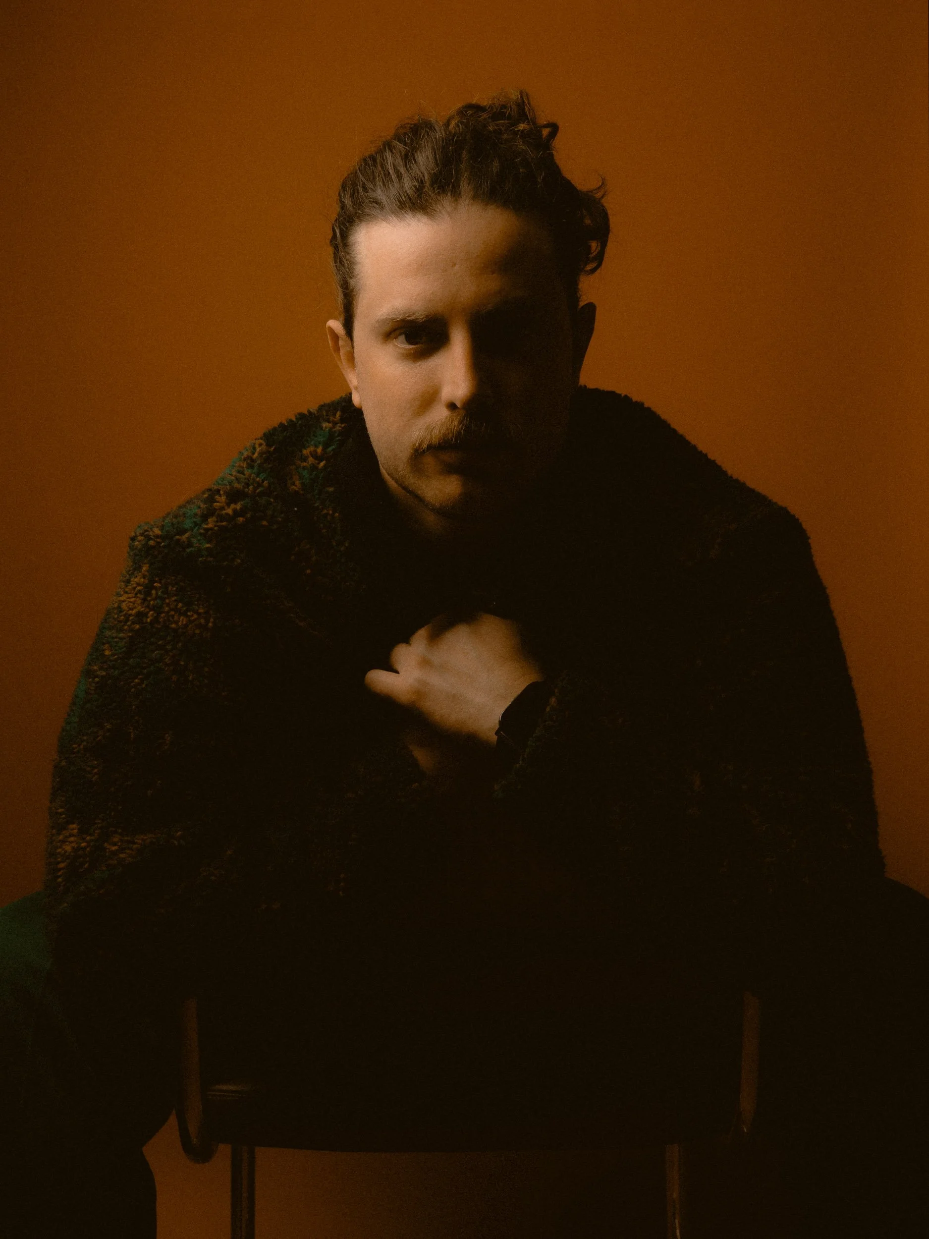 A man with dark hair and a mustache sitting on a chair with a warm brown background. He is wearing a textured dark coat and looking directly at the camera with a serious expression. Personal Branding Photography and Headshots in London.