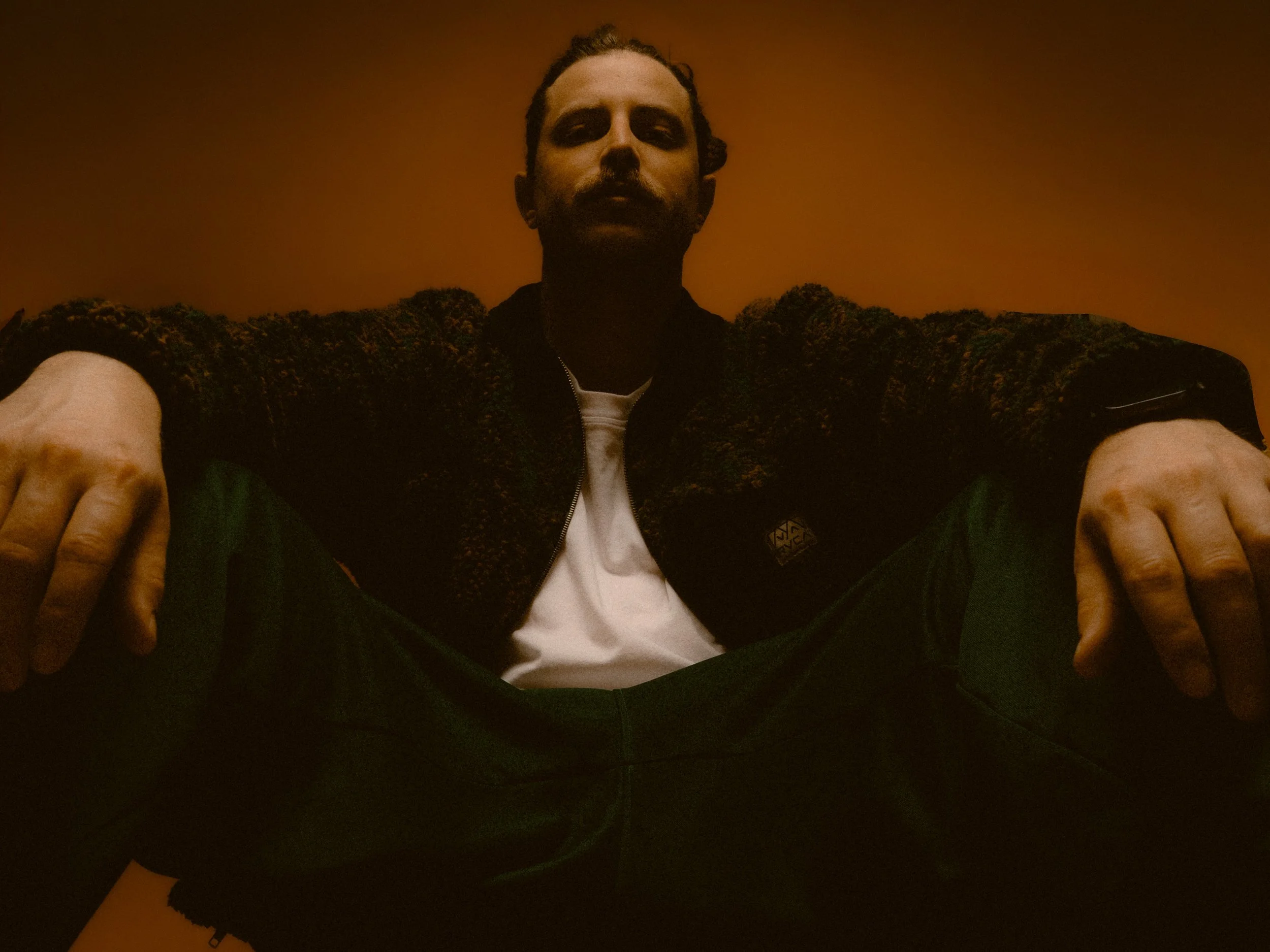 Low-angle photo of a man sitting with legs apart, wearing a black jacket, white T-shirt, and green pants, against an orange background. Personal Branding Photography and Headshots in London.
