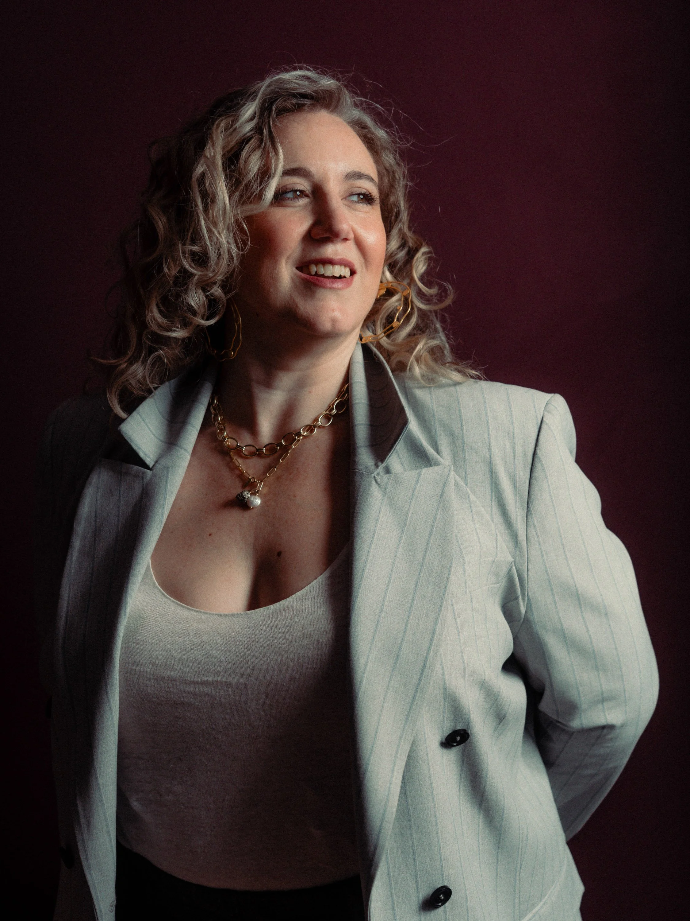 A woman with curly blonde hair wearing a light striped blazer, a white top, and gold jewelry, standing against a dark background. Personal Branding Photography and Headshots in London.