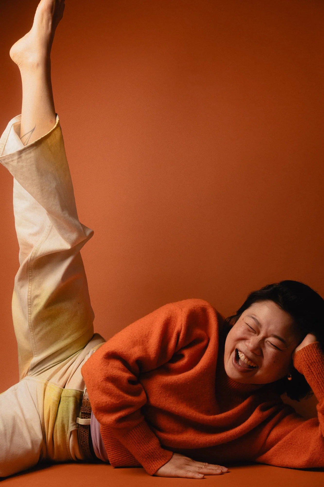 A woman laughing while lying on an orange surface against an orange background, wearing an orange sweater and beige pants.