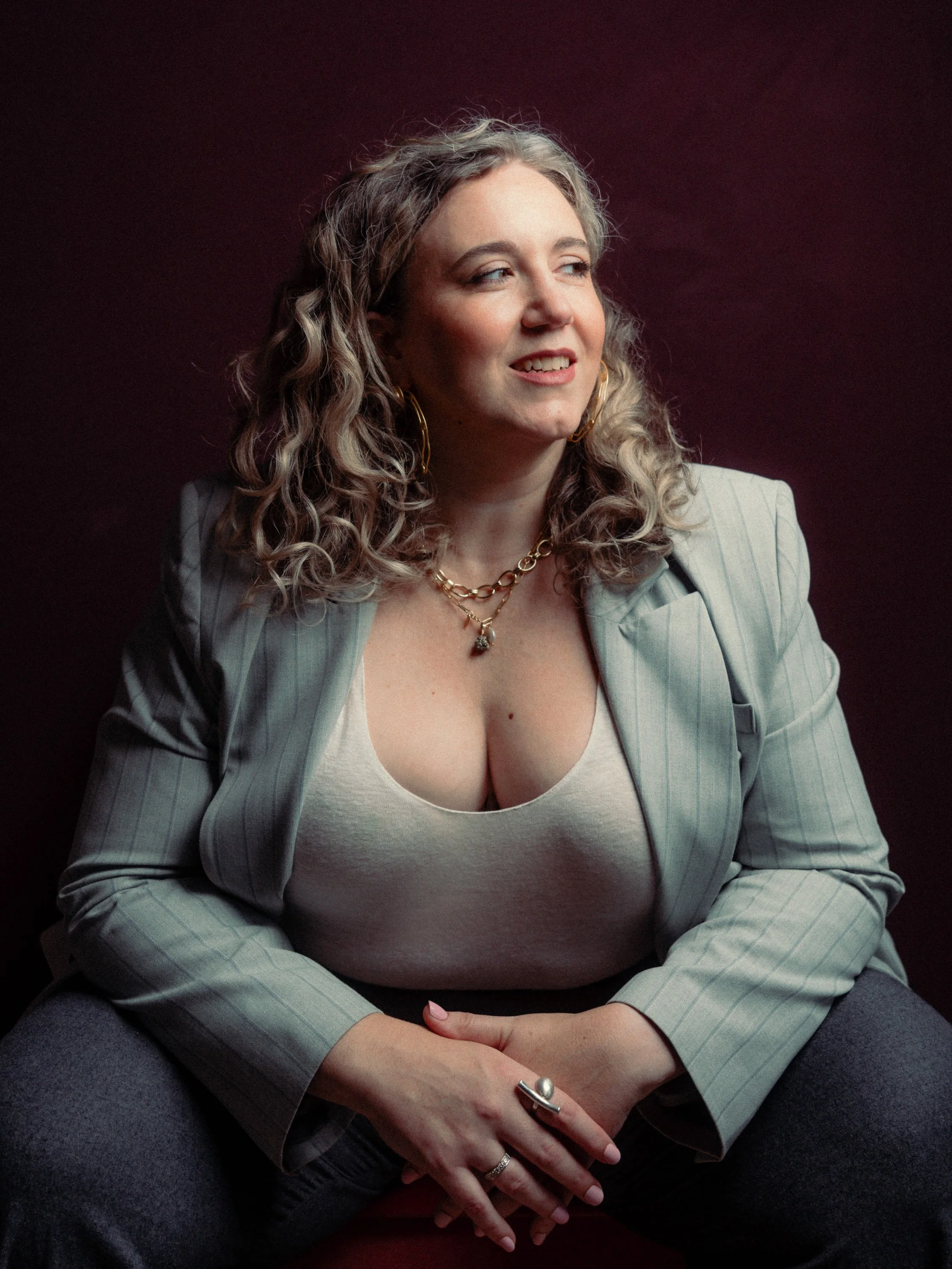 A woman with curly blonde hair wearing a light-colored blazer, a white top, and gold jewelry, sitting against a dark background. Personal Branding Photography and Headshots in London.