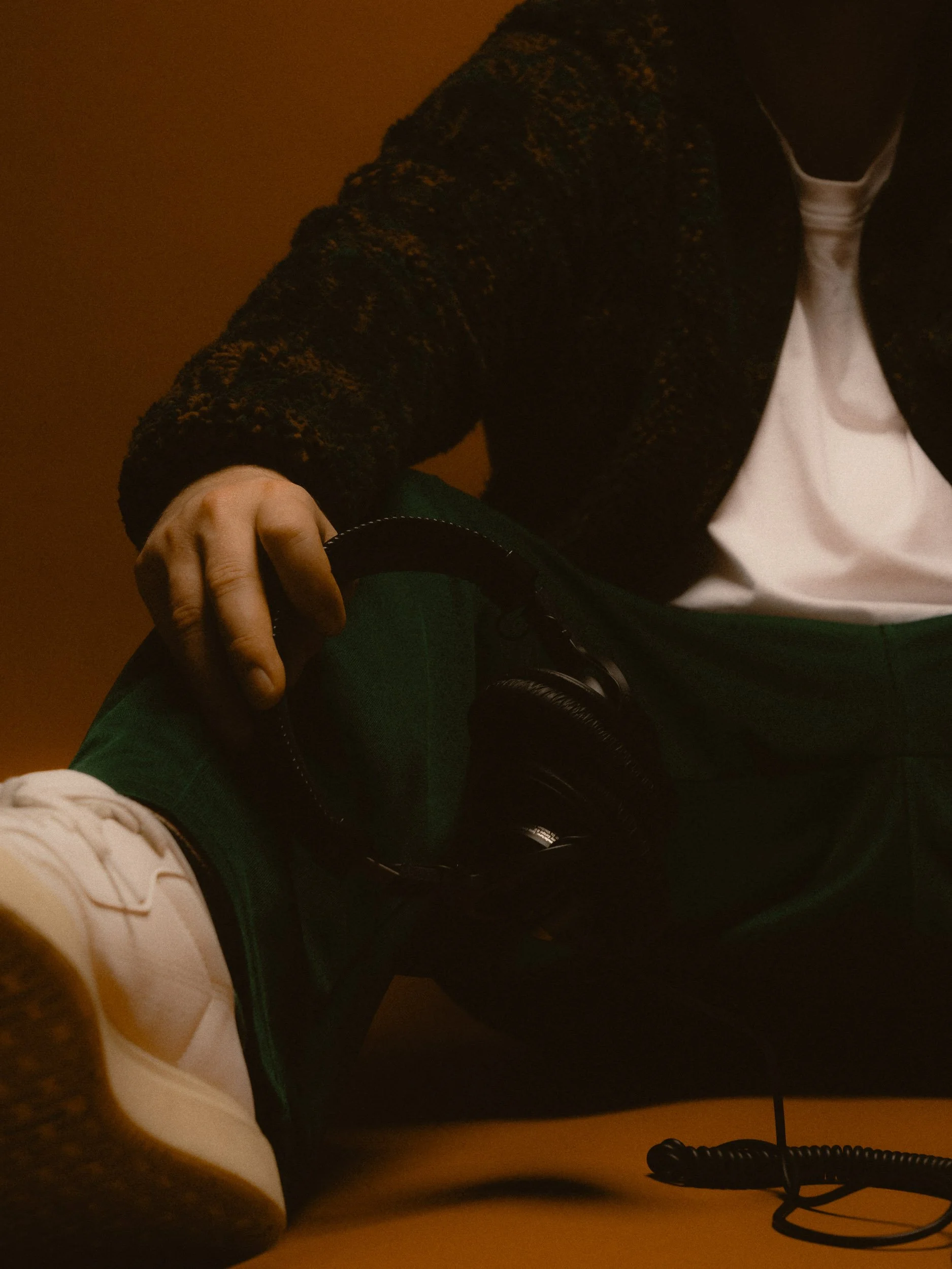 Person sitting with their legs crossed, wearing white sneakers, green plaid pants, a black patterned jacket, white shirt, and holding black headphones. Personal Branding Photography and Headshots in London.