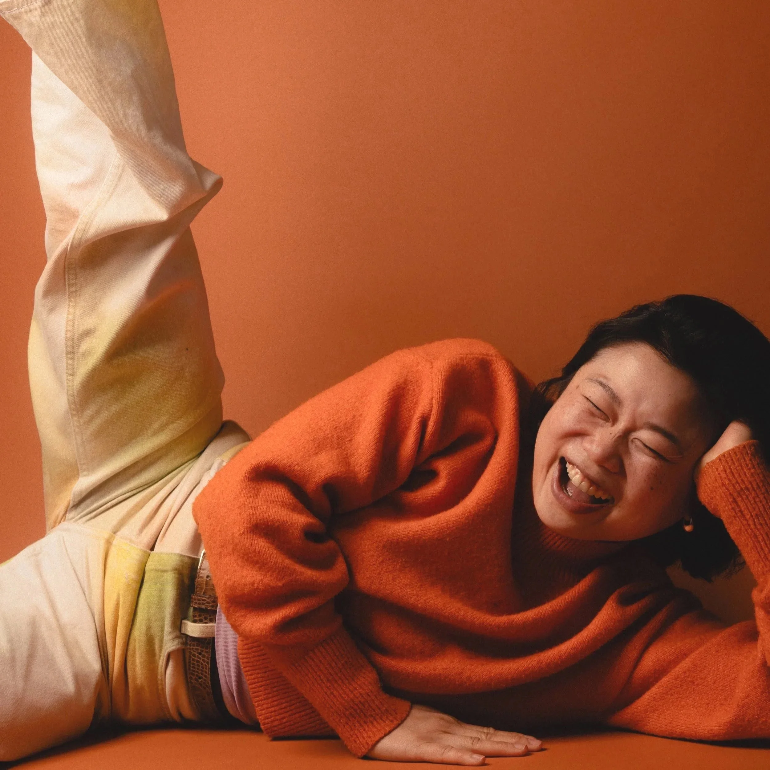 A woman laughing while lying on an orange surface against an orange background, wearing an orange sweater and beige pants.