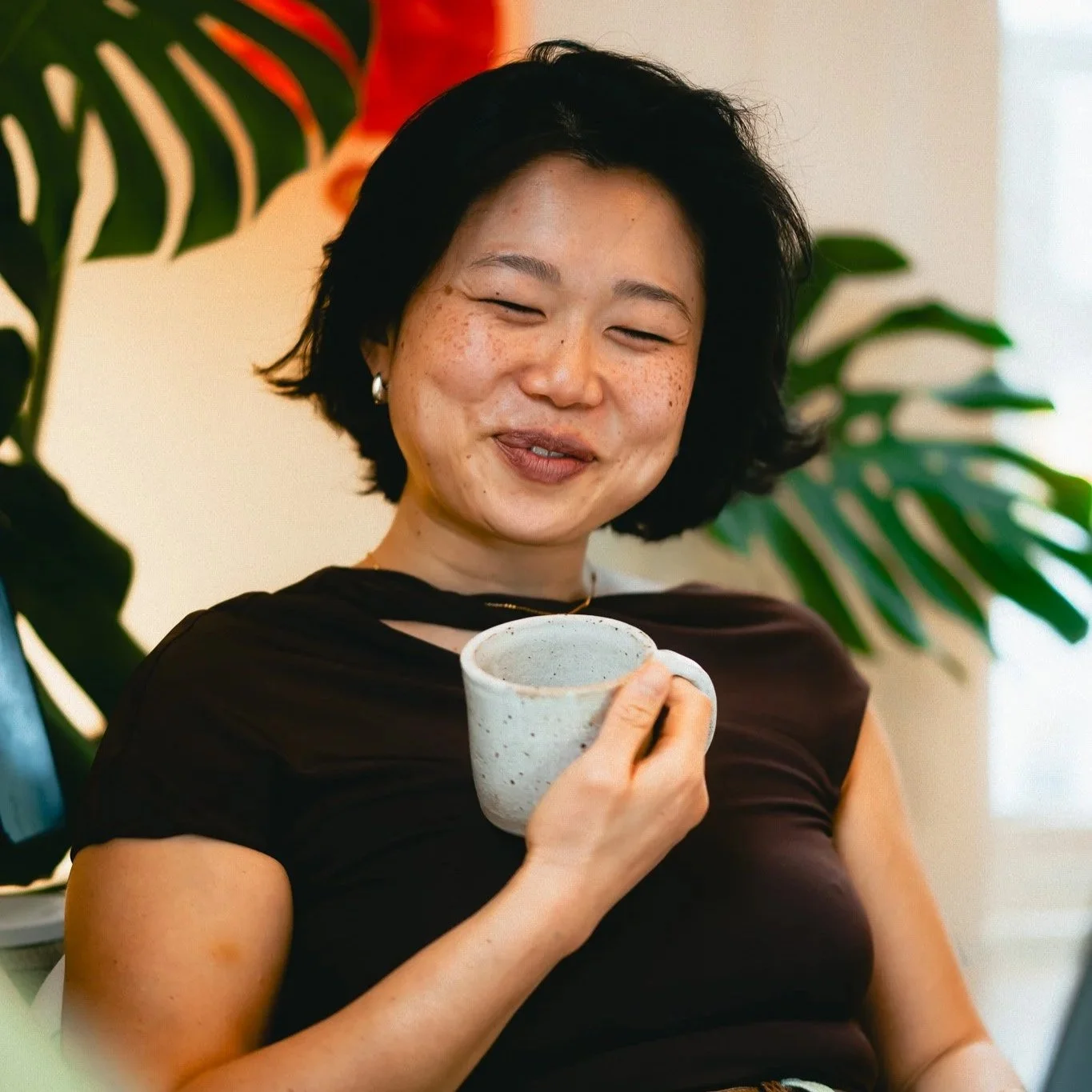 A woman with short black hair and freckles is smiling and holding a speckled ceramic mug, sitting indoors with large green plants in the background.
