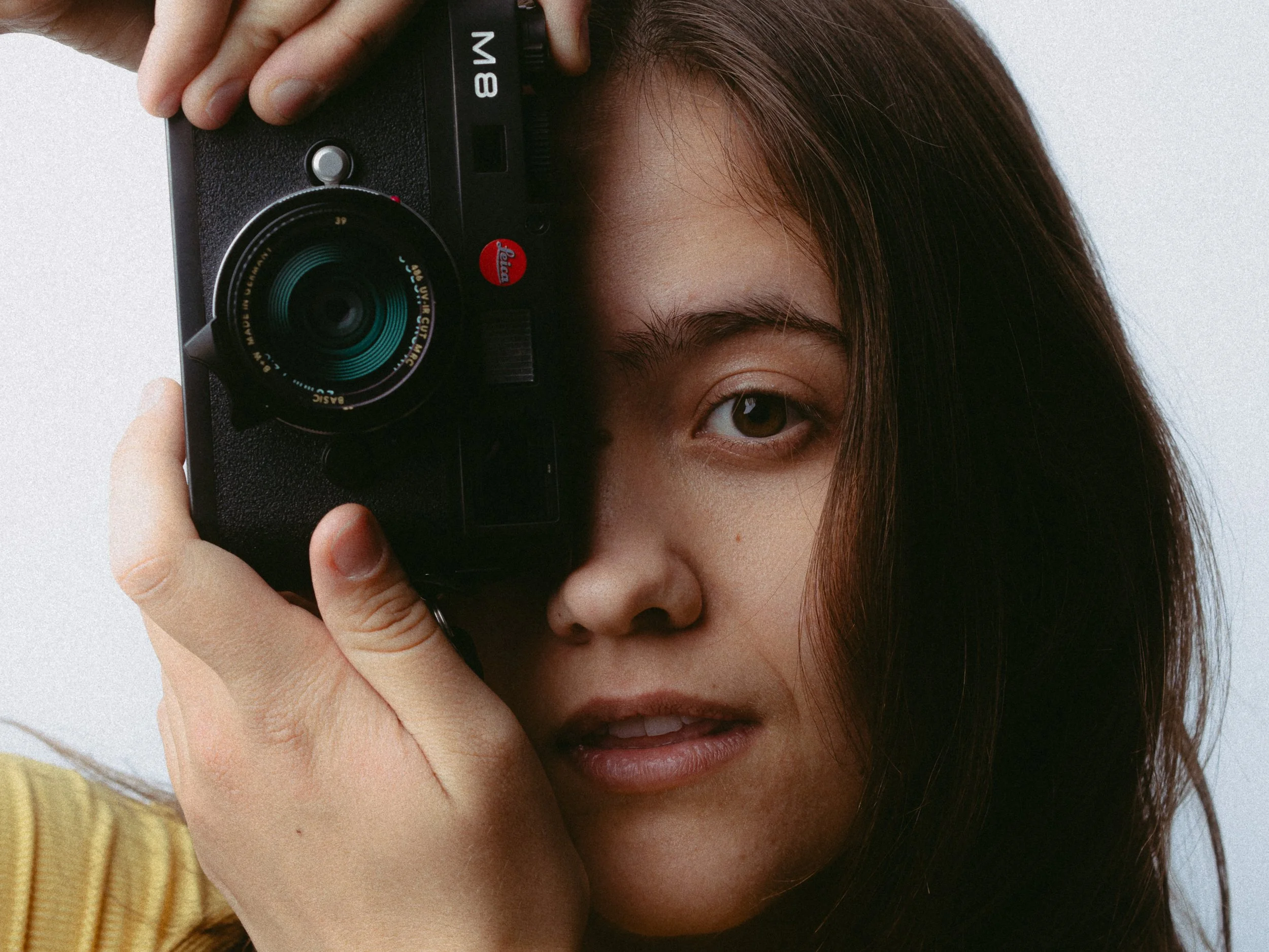 A woman holding a camera up to her face, with her right eye peeking behind the camera, and brown hair falling to her shoulder. Personal Branding Photography and Headshots in London.
