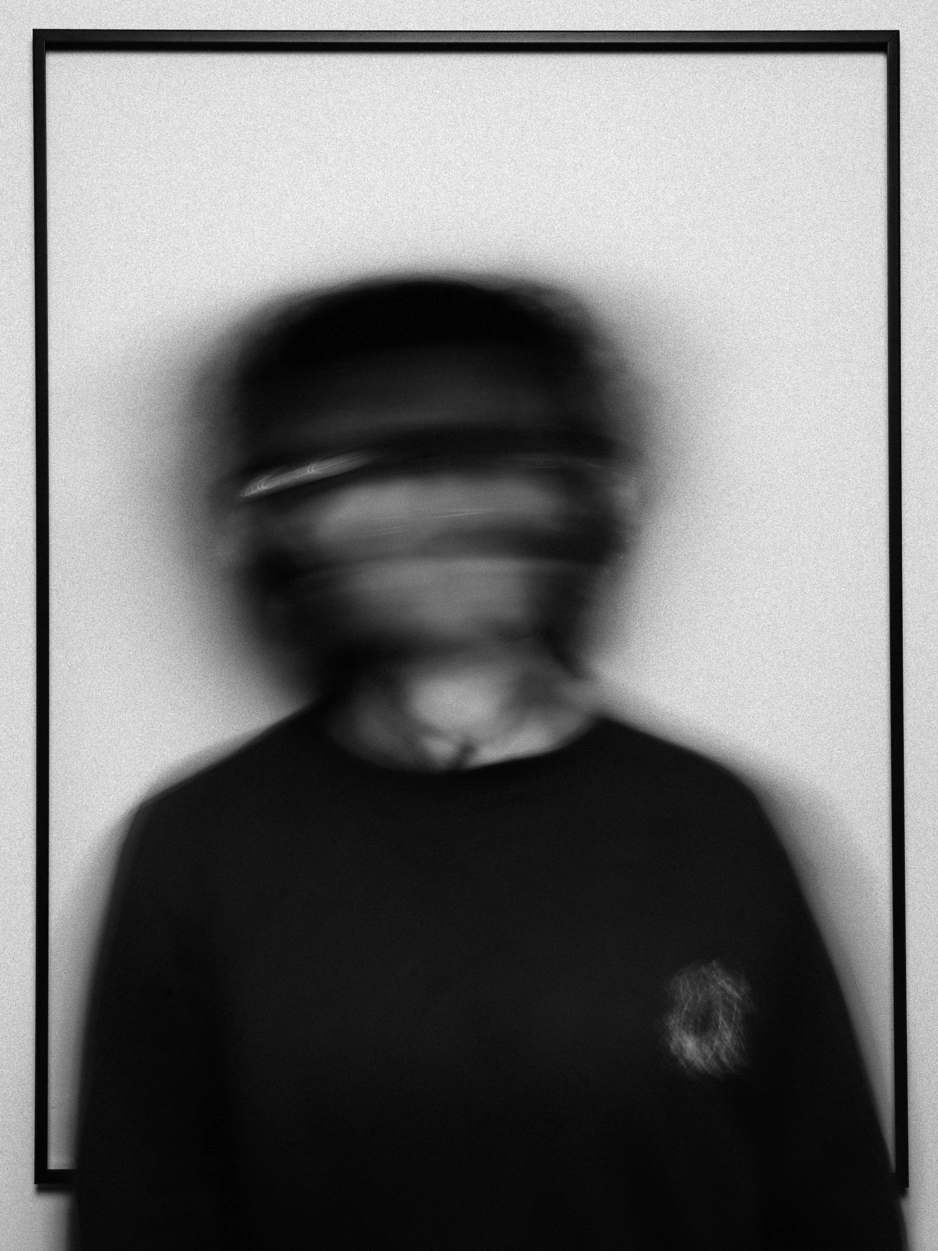 A blurry black-and-white photo of a person with short hair, wearing glasses and a dark shirt, standing in front of a plain wall. Personal Branding Photography and Headshots in London.