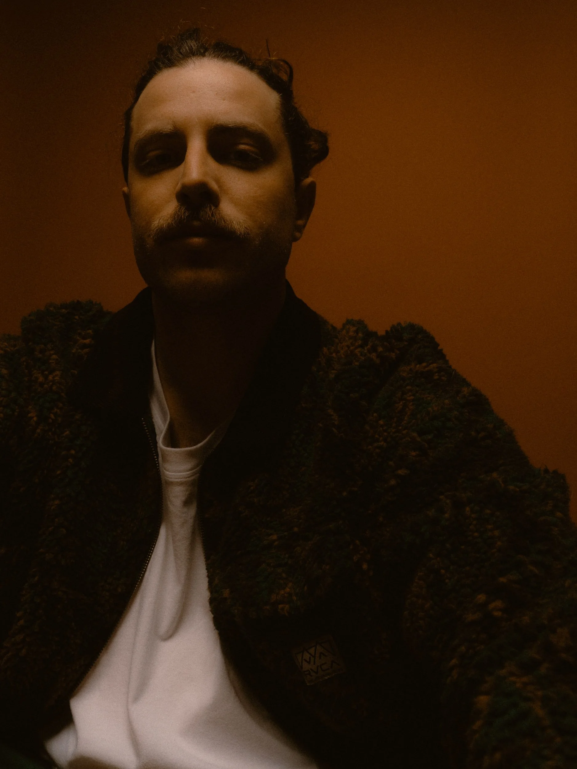 A man with a mustache and curly hair, wearing a patterned jacket and a white shirt, sits against a dark background with a warm, orange hue. Personal Branding Photography and Headshots in London.