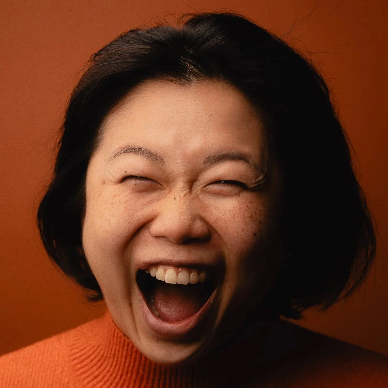 A woman with short black hair and freckles, wearing an orange sweater, laughing with her mouth open and eyes closed against a warm orange background.