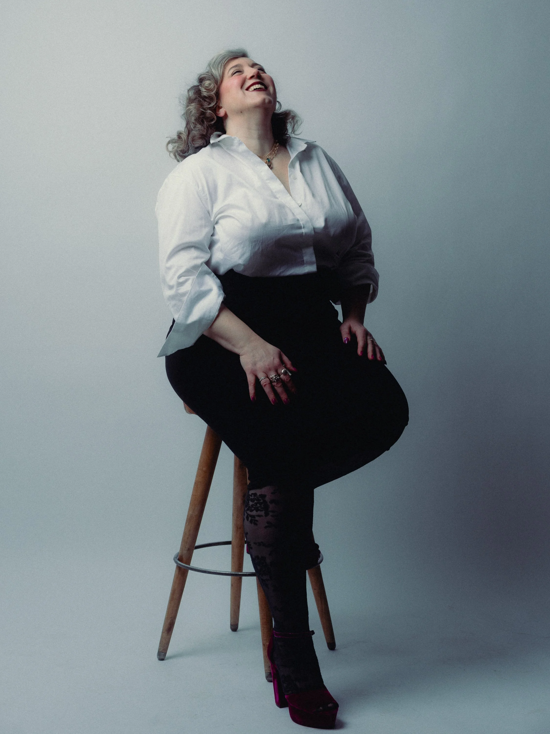 A woman with gray curly hair smiling while sitting on a wooden stool against a plain gray background. Personal Branding Photography and Headshots in London.