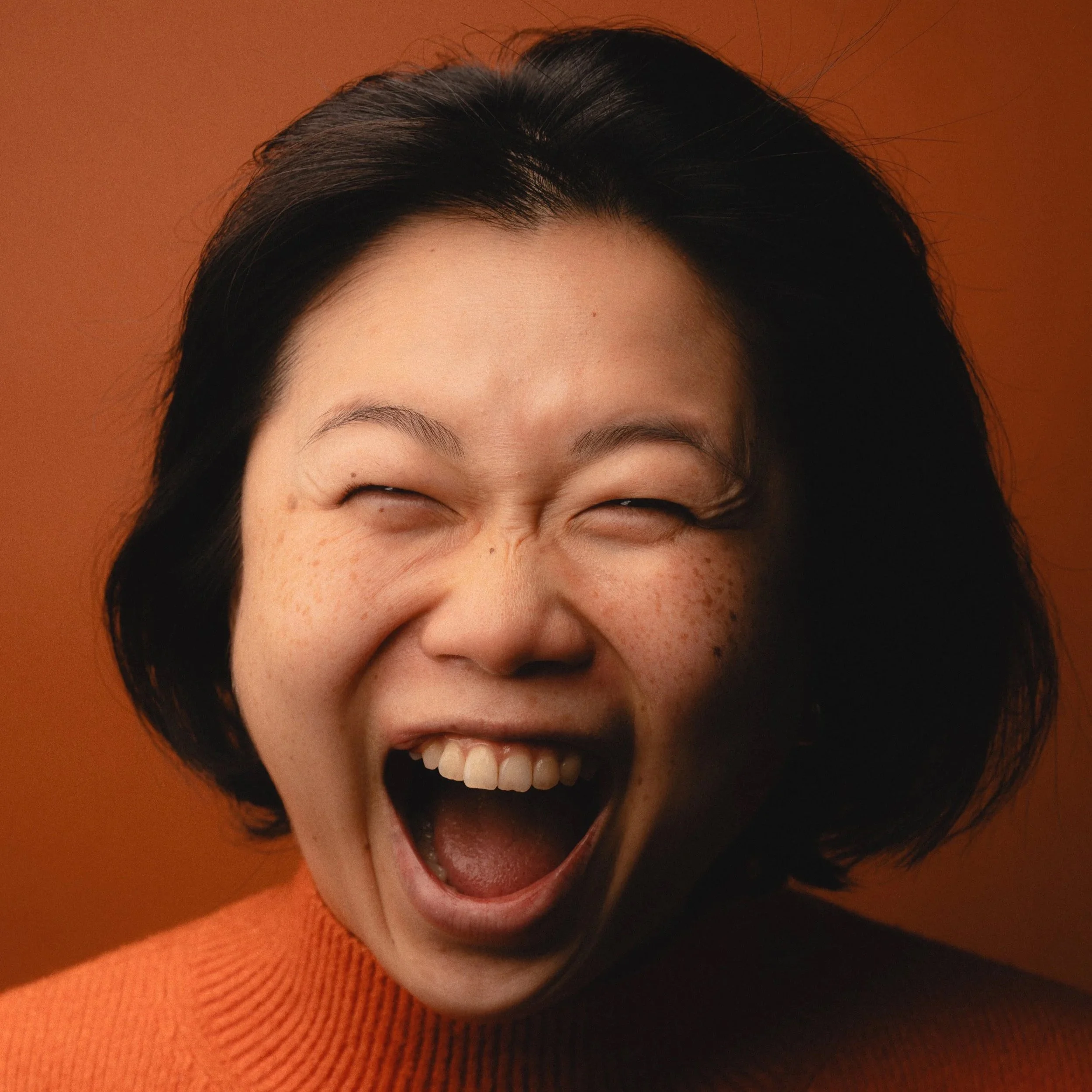 A woman with short black hair and freckles, wearing an orange sweater, laughing with her mouth open and eyes closed against a warm orange background.