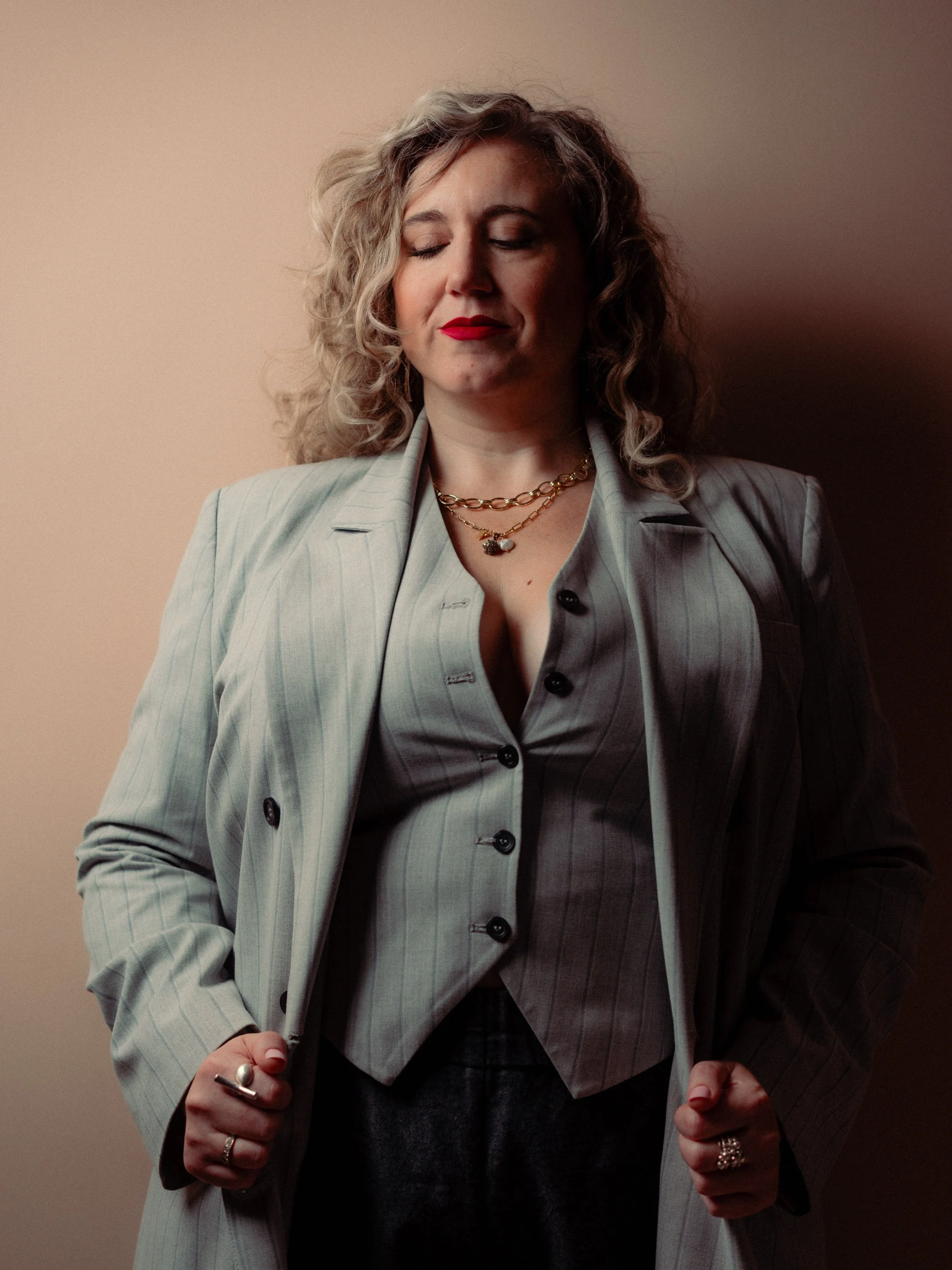 A woman with curly blonde hair and red lipstick wearing a light gray pinstripe suit with a blazer and vest, accessorized with layered gold necklaces and rings. Personal Branding Photography and Headshots in London.