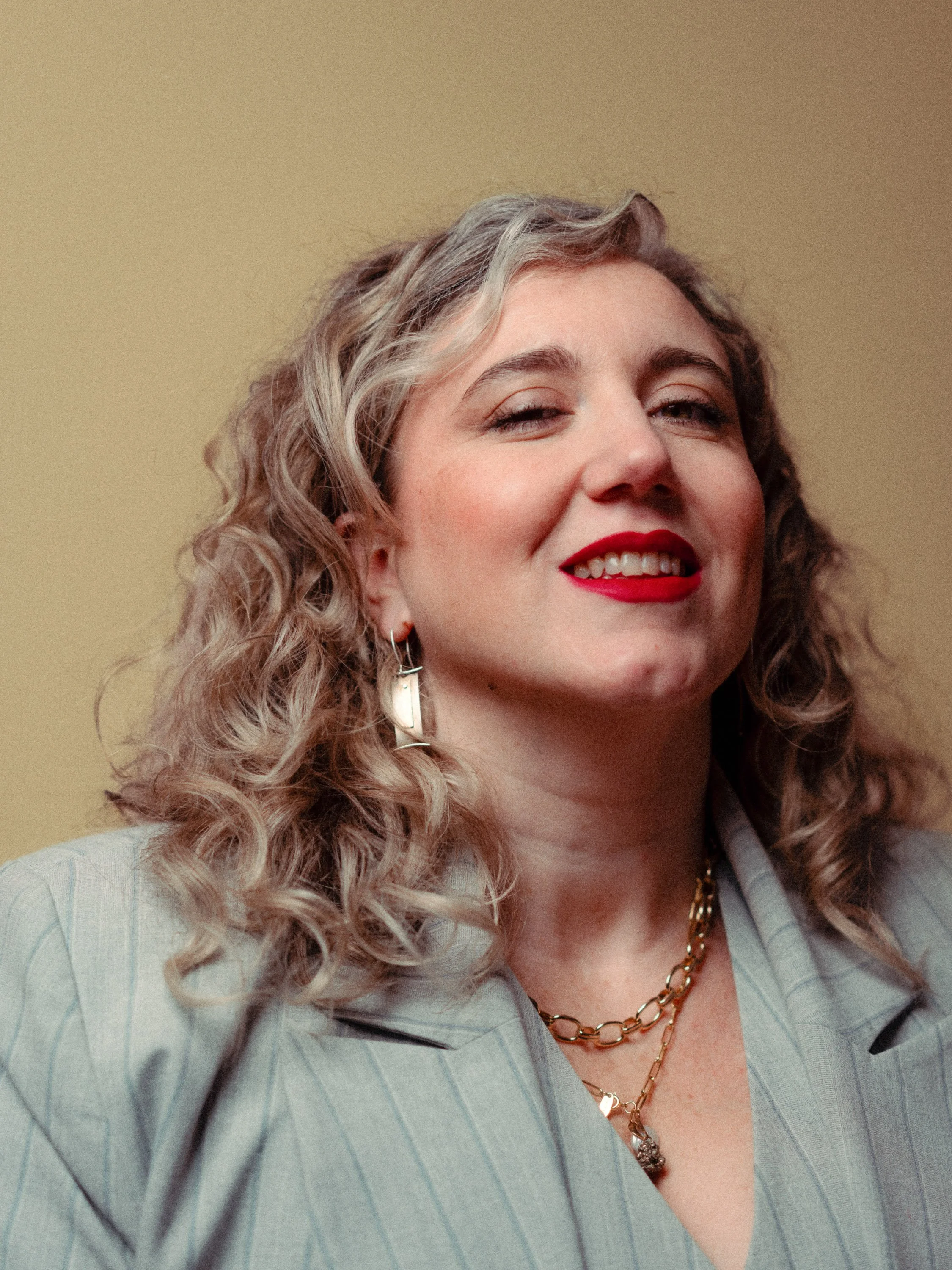 A woman with curly blonde hair, red lipstick, and a confident smile, wearing jewelry and a gray blazer against a beige background. Personal Branding Photography and Headshots in London.