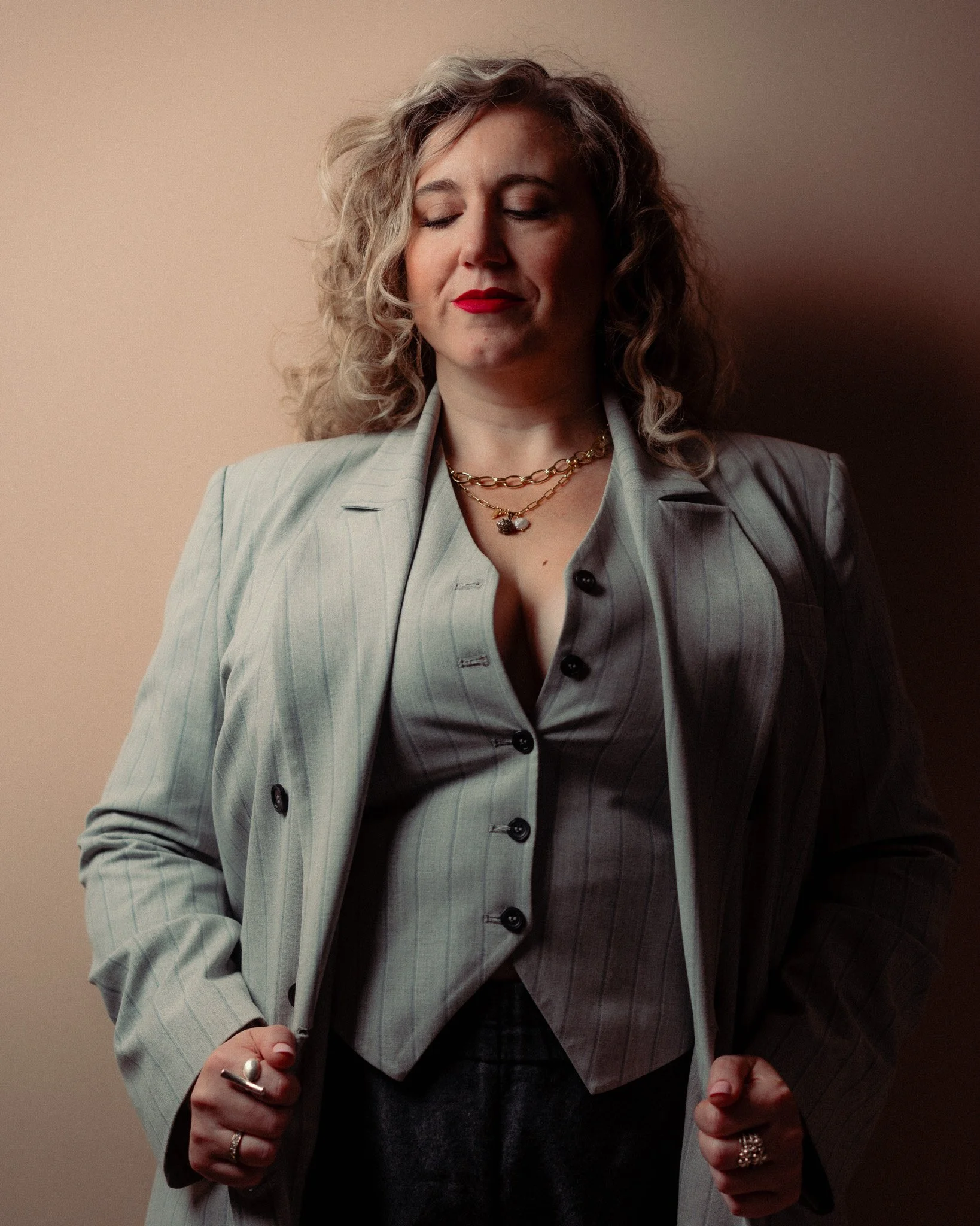A woman with curly blonde hair and red lipstick wearing a light gray pinstripe blazer and vest, layered gold necklaces, and rings, standing against a beige background with her eyes closed.