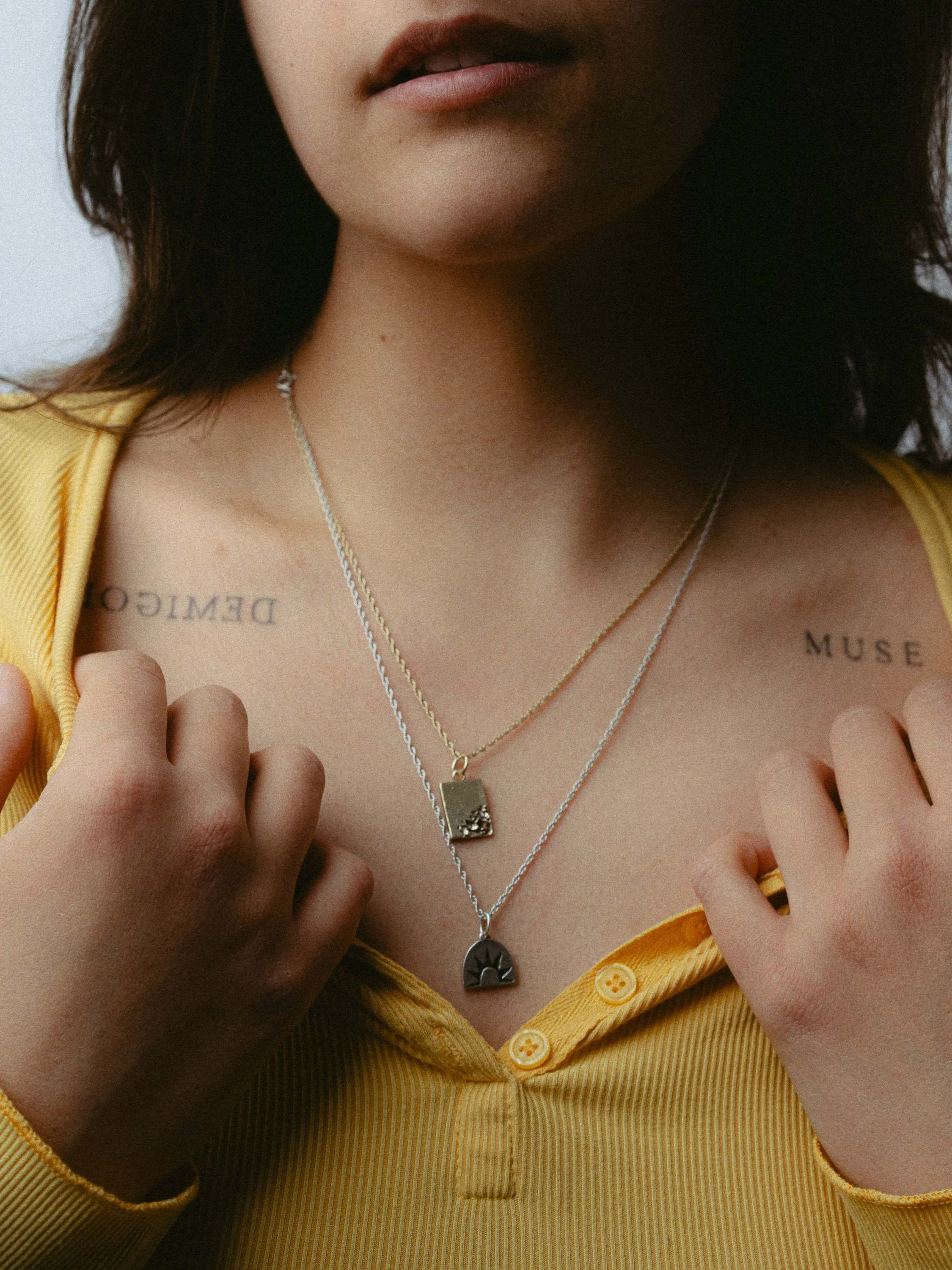 A woman in a yellow button-up shirt shows two layered necklaces with rectangular and semi-circular pendants, and tattoos on her collarbone reading 'DEMOCRACY' and 'MUSE'. Personal Branding Photography and Headshots in London.