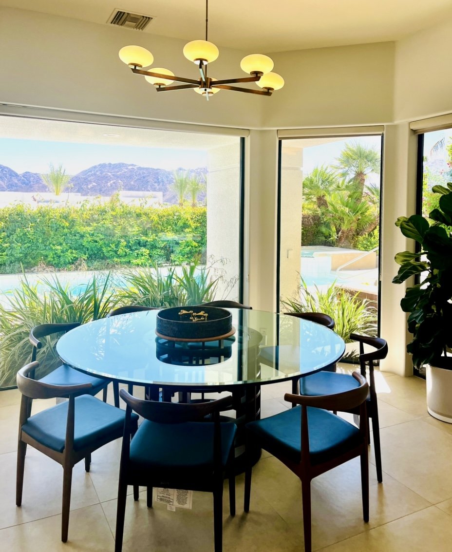 Dining room with a round glass table surrounded by six dark wood chairs, a modern chandelier on the ceiling, large windows showing green plants and mountains, and a potted plant in the corner.